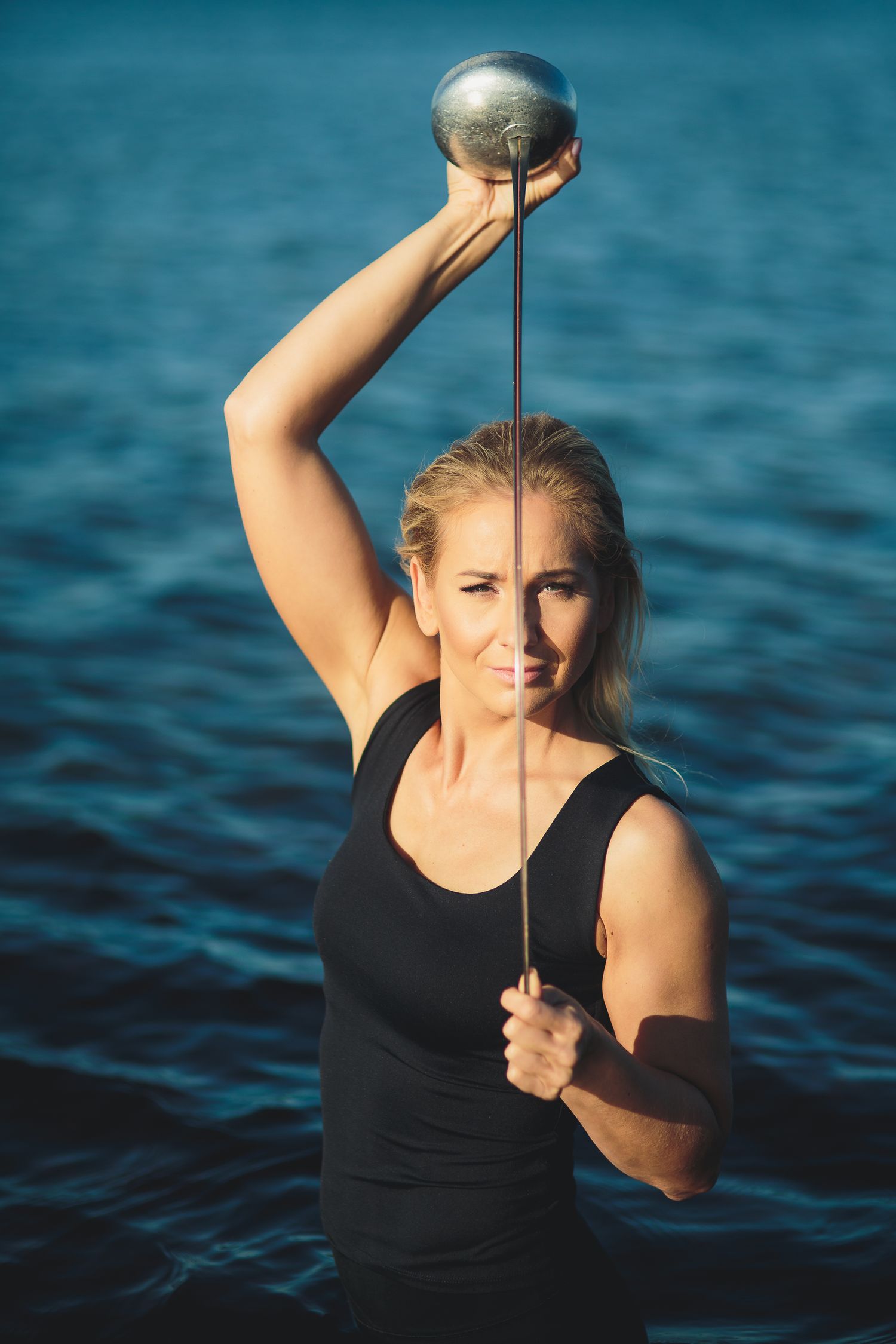 Athletic blonde woman lifting a kettlebell overhead with blue water in the background.