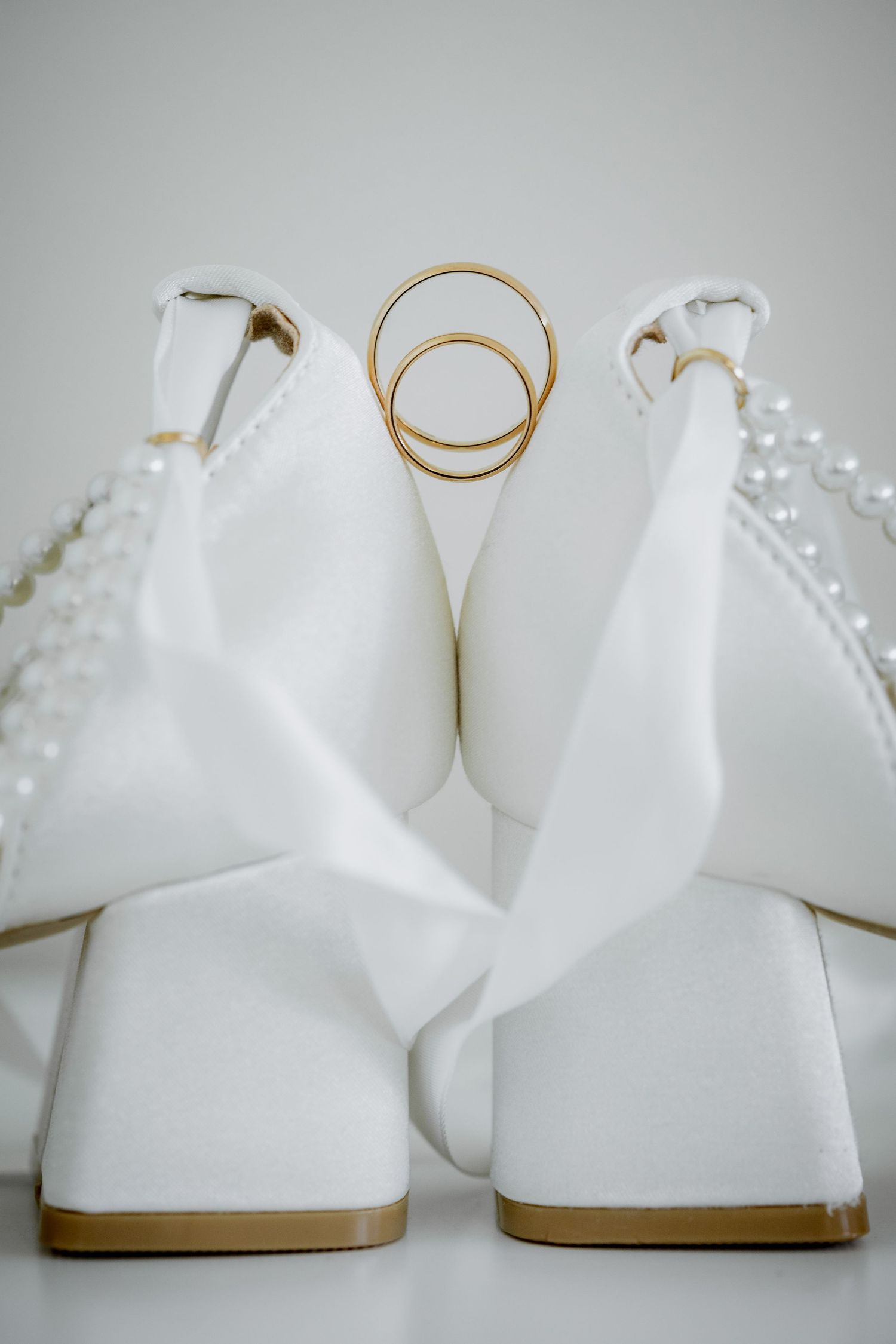 White bridal heels with pearl embellishments and two gold wedding rings nestled between them on a white background.