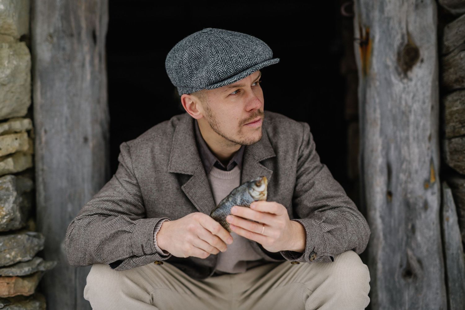Man in vintage tweed jacket and flat cap holding a fish while sitting in a rustic stone doorway.