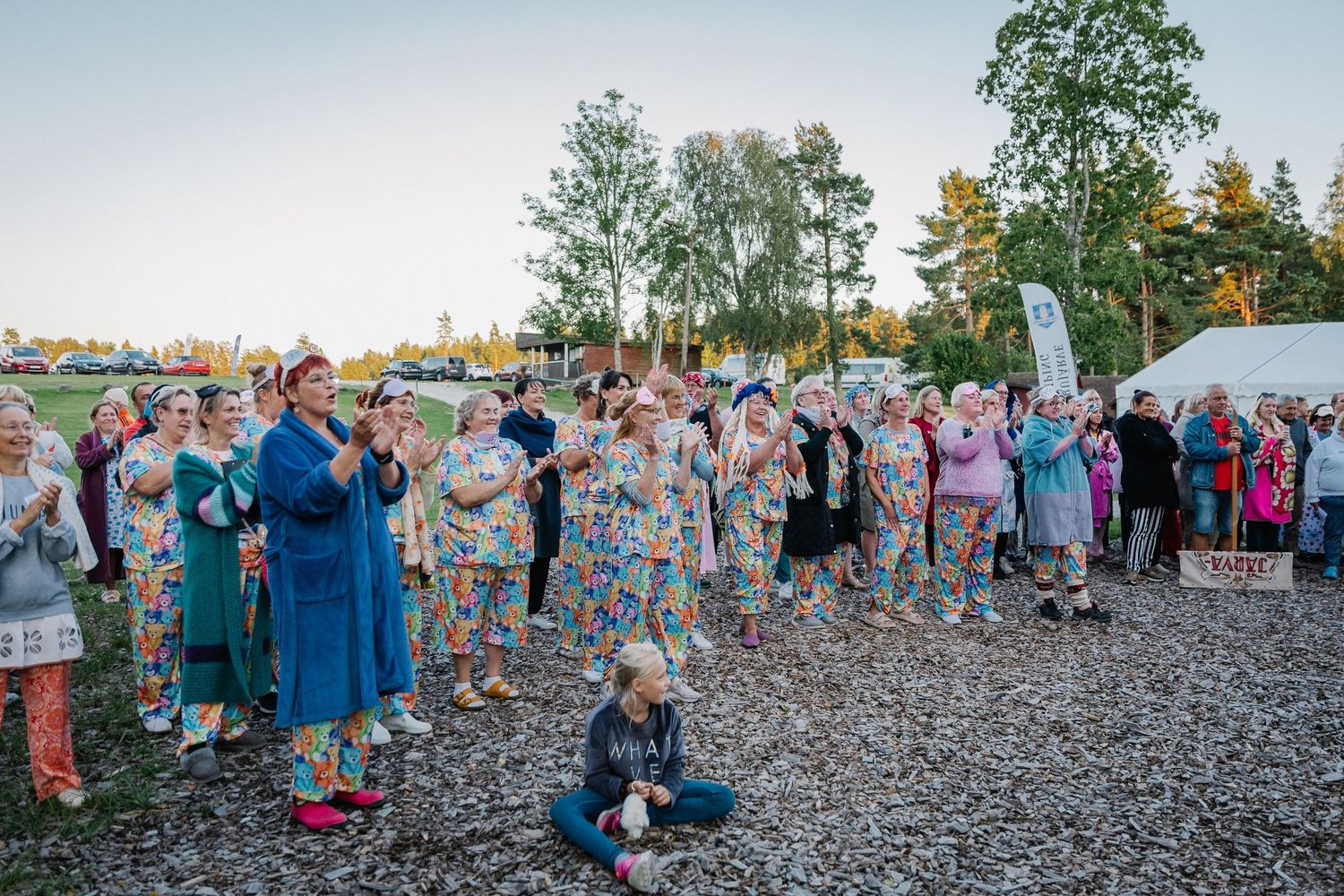 Large group of people wearing colorful patterned jumpsuits gathered outdoors at an evening event near trees.