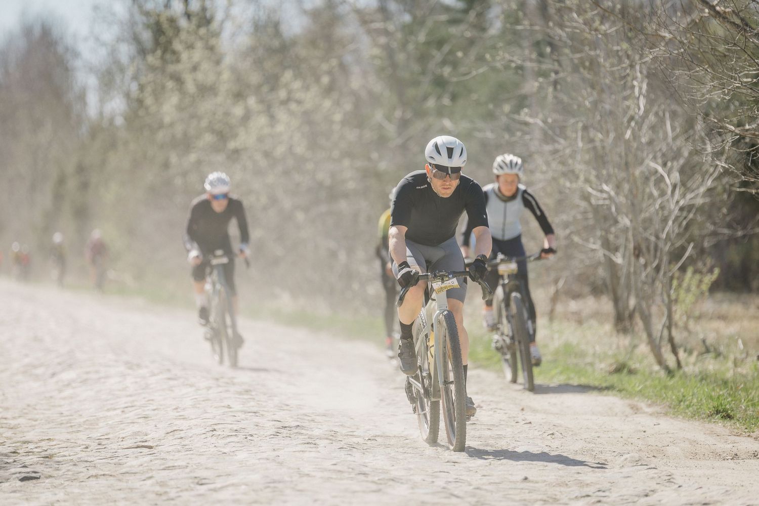 Three cyclists riding mountain bikes on a dusty gravel path through a sunny forest trail.