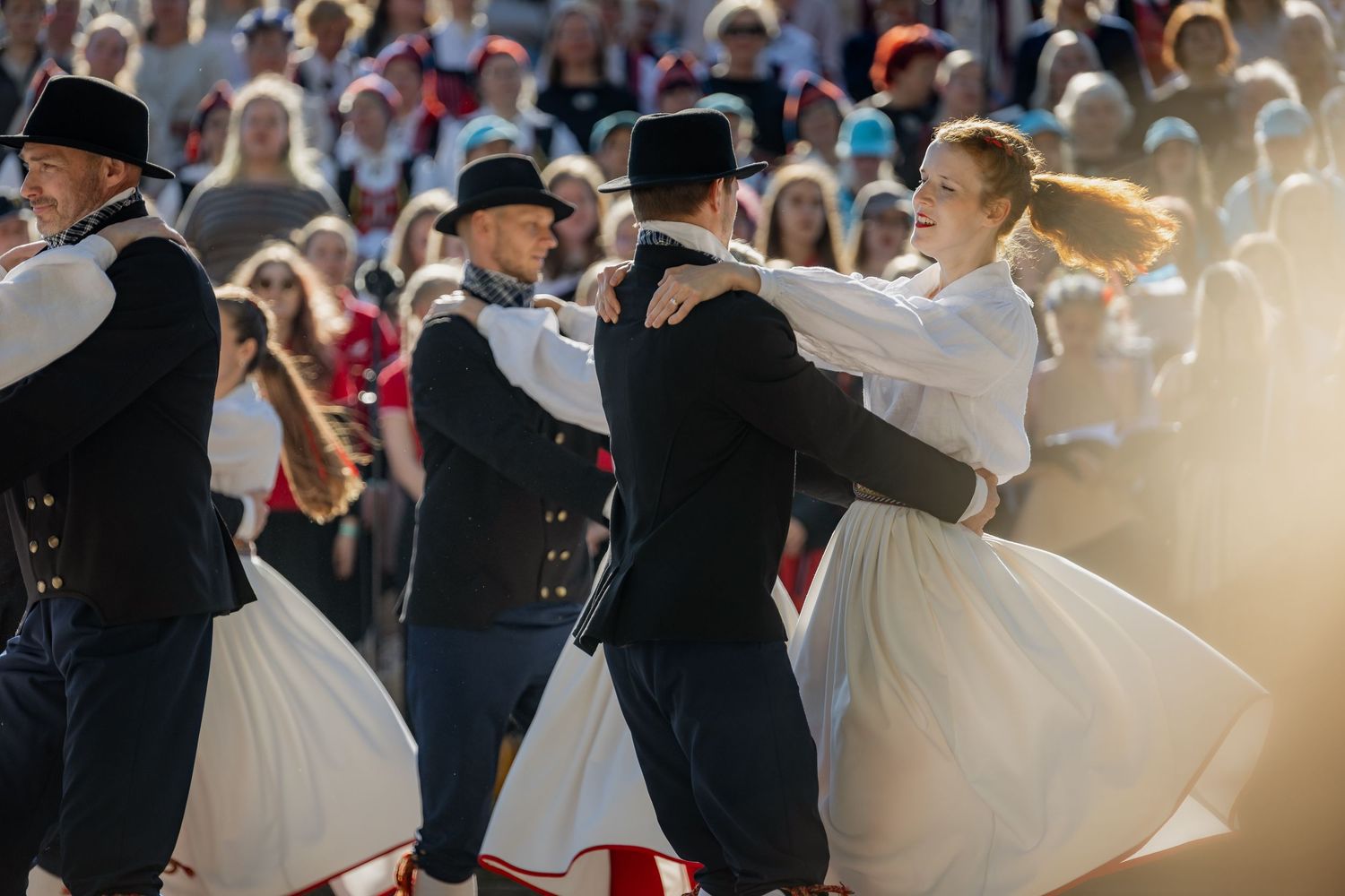 Traditional folk dancers in black and white costumes perform at an outdoor festival before a large crowd.