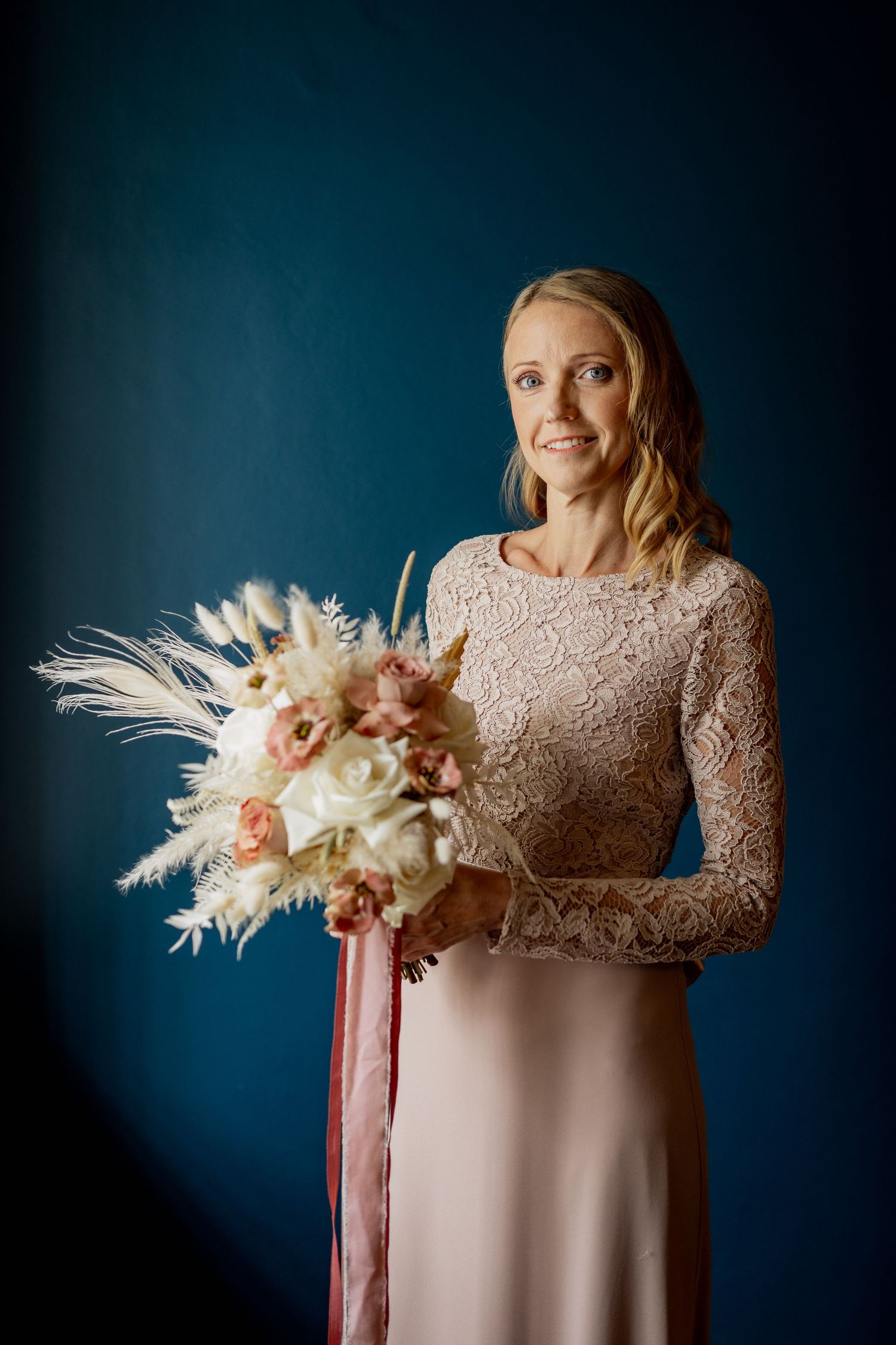 Smiling bride in lace dress holding boho bouquet with pampas grass and pink roses against blue backdrop.