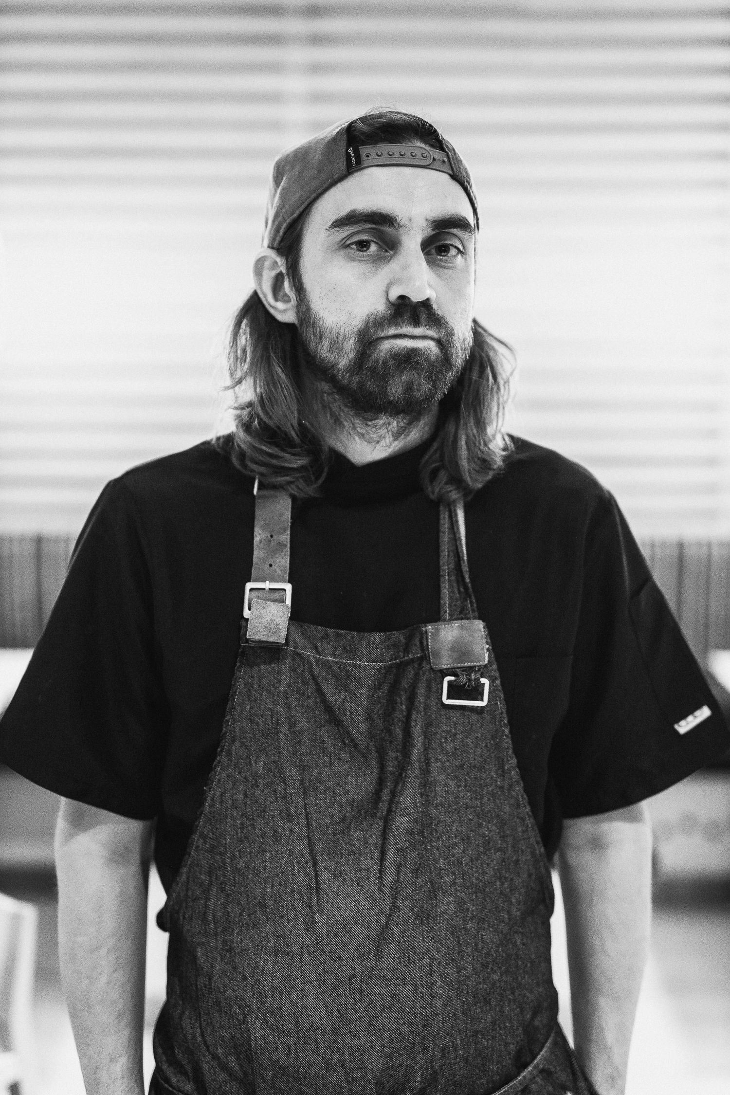 Black and white portrait of bearded man with long hair wearing apron and backward cap indoors.