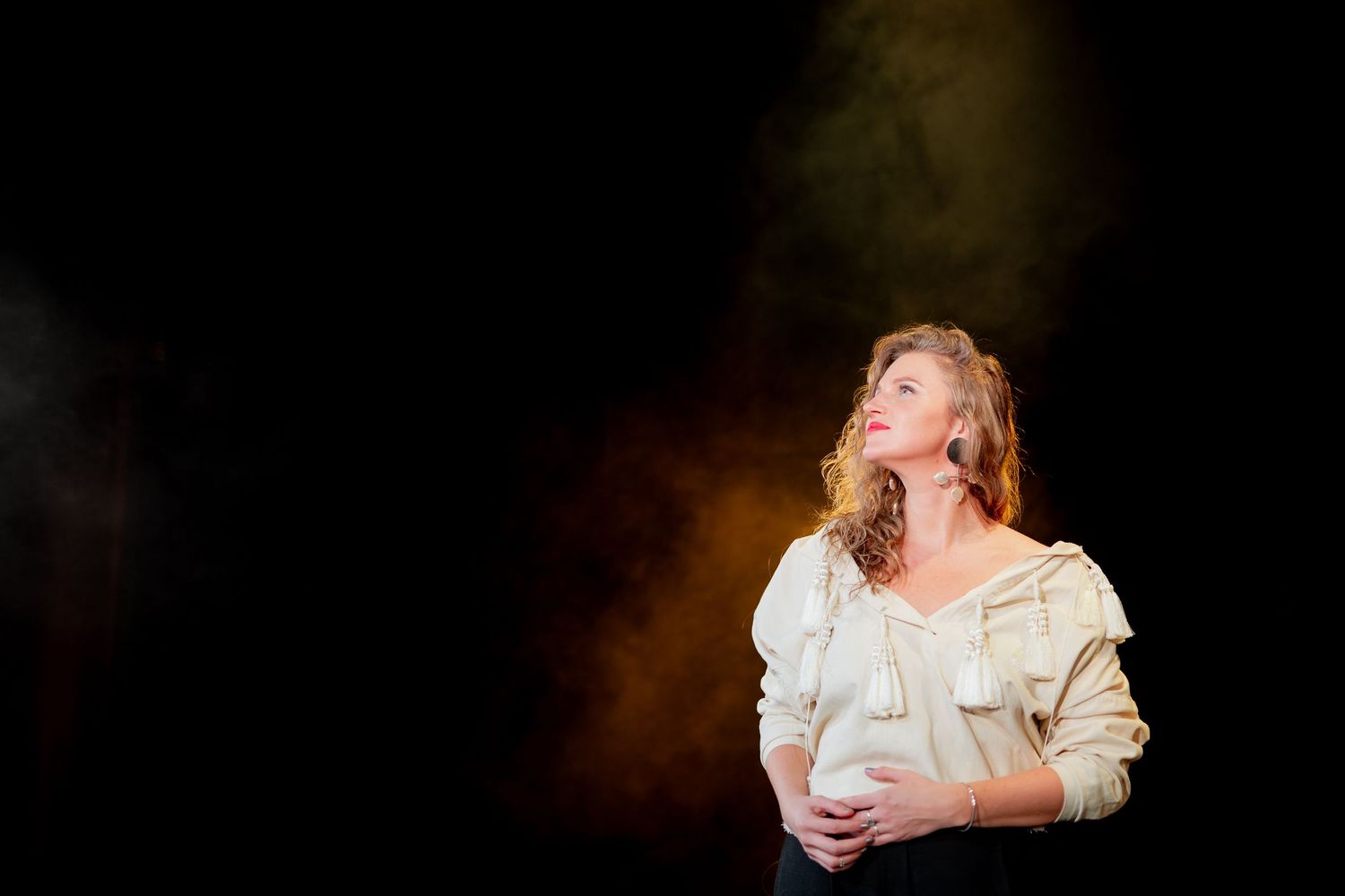 Young woman in a white blouse looking upward dramatically against a dark smoky stage background.