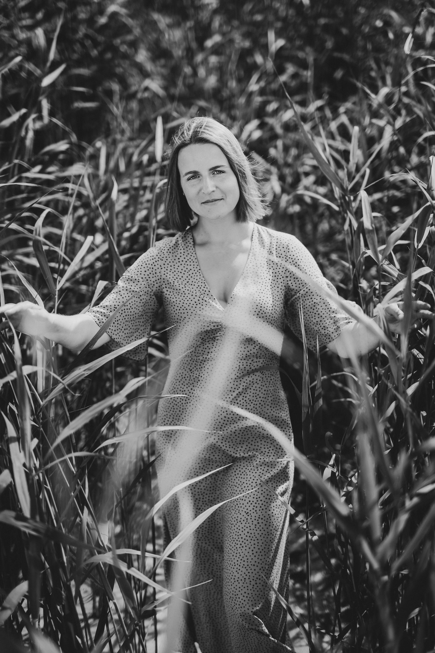 Woman in spotted dress parts tall grass reeds with hands in artistic black and white portrait.