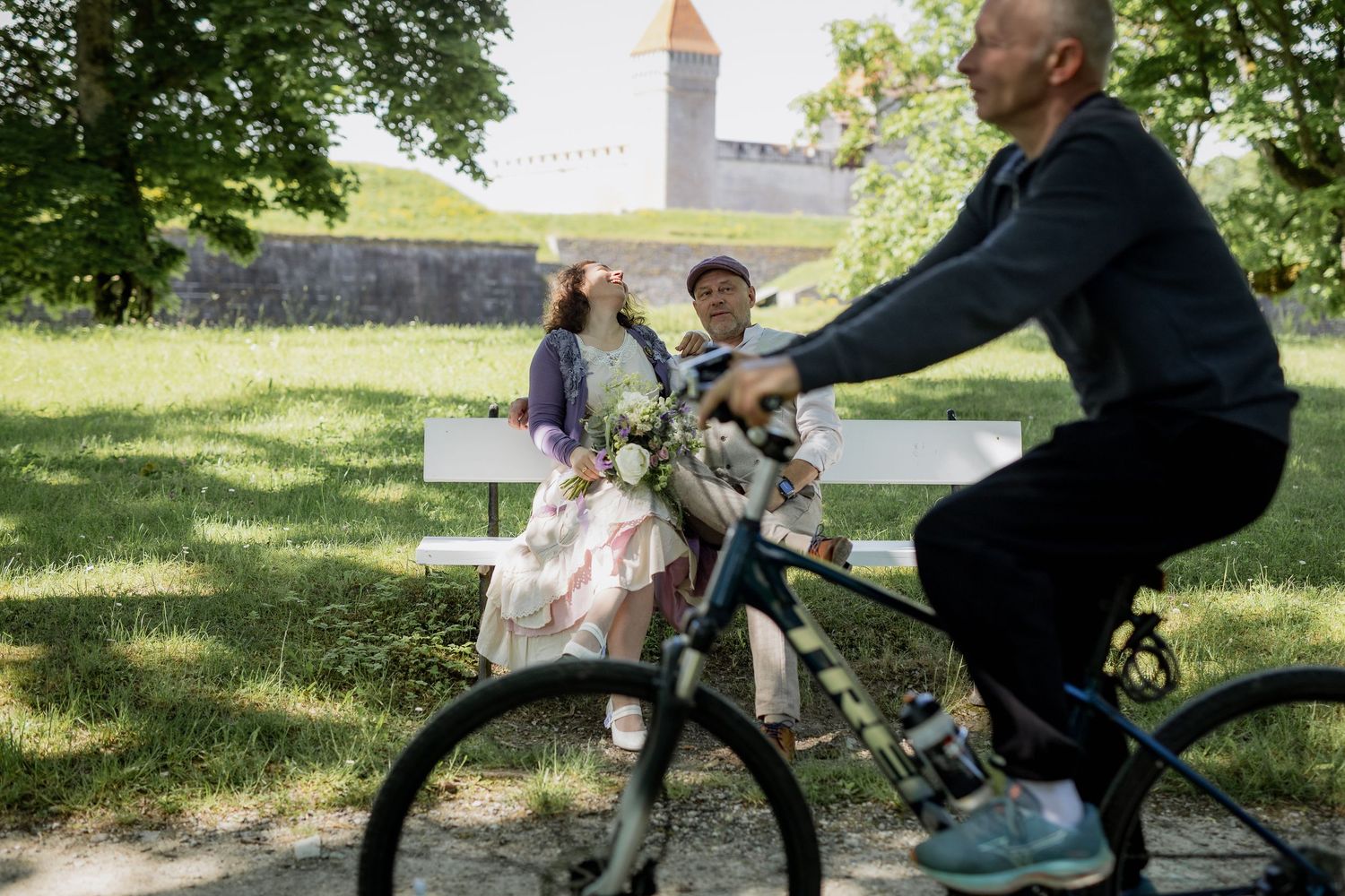A man rides a bicycle past an elderly couple sitting on a white park bench near a historic stone castle.