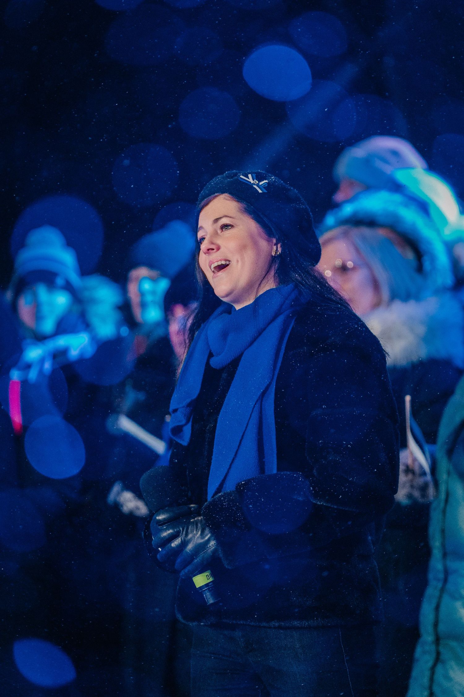 Woman in blue scarf and black coat singing outdoors at a winter night event with bokeh lights in background.