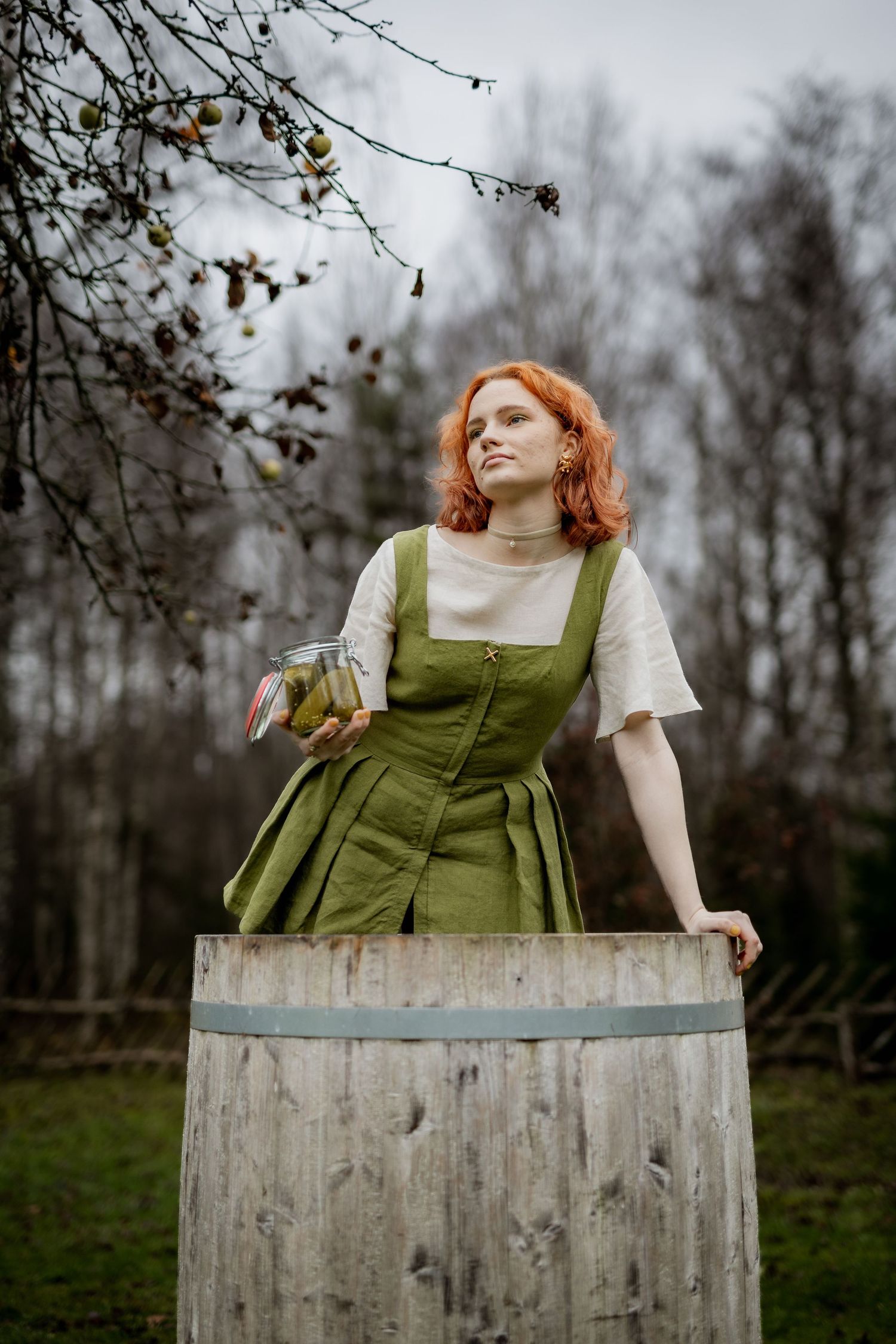 Red-haired woman in green linen dress leaning on wooden barrel outdoors in autumn garden setting.