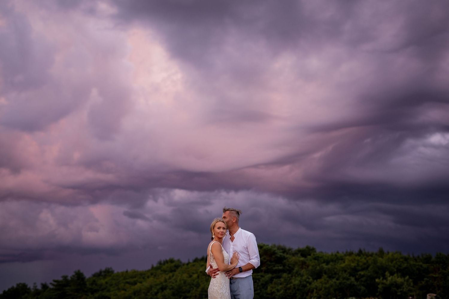 Couple embraces under dramatic purple storm clouds at sunset in an outdoor wedding portrait.