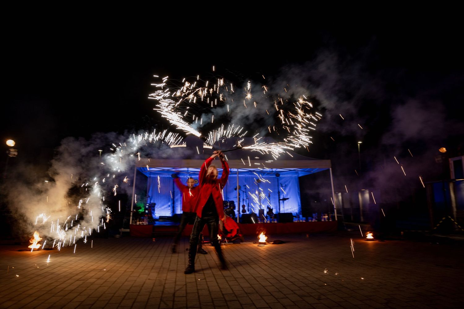 Performer in red costume dances with sparkling fireworks display at night outdoor event with blue lit stage backdrop.