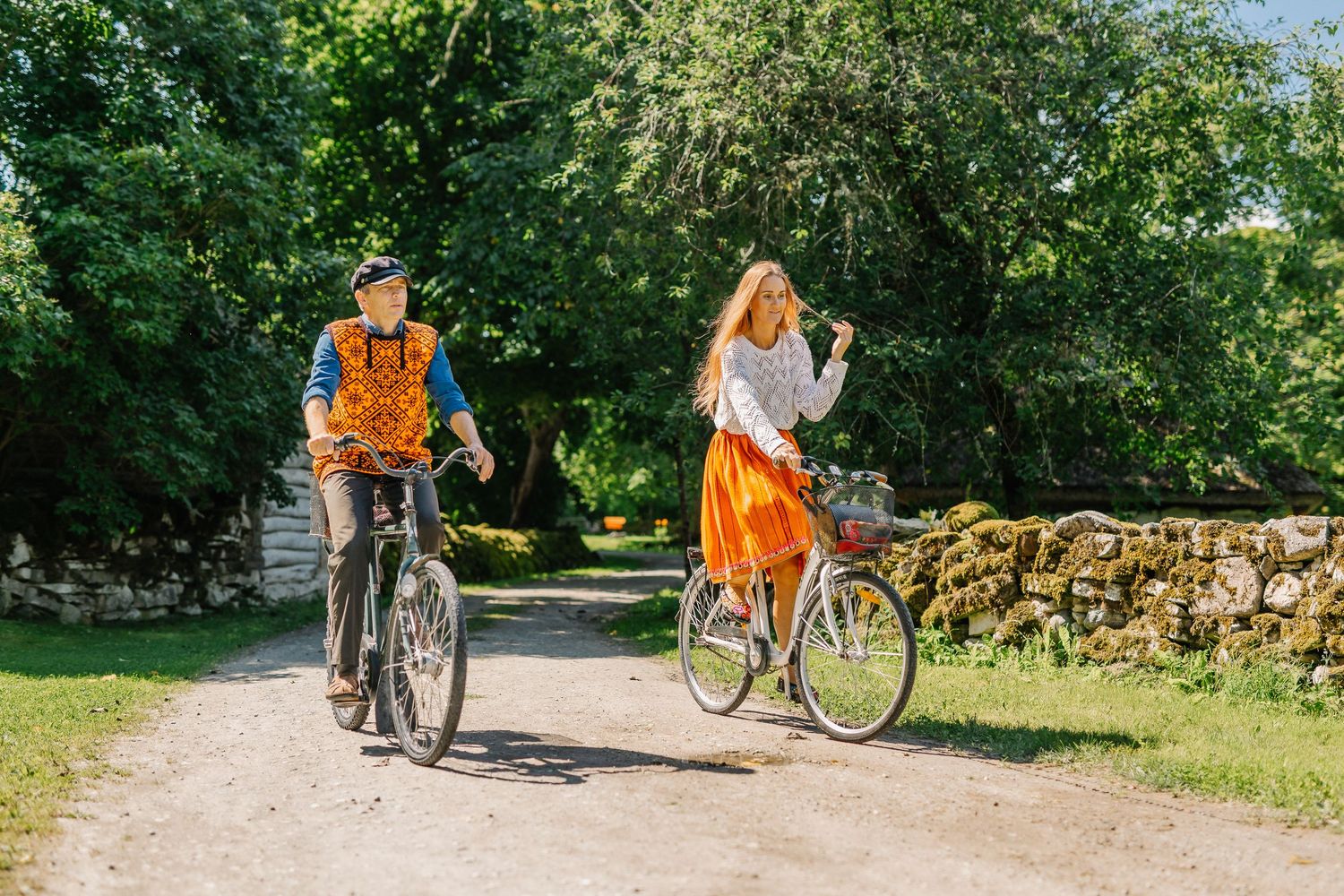 Couple riding bicycles on a scenic gravel path surrounded by lush green trees and a stone wall on a sunny day.