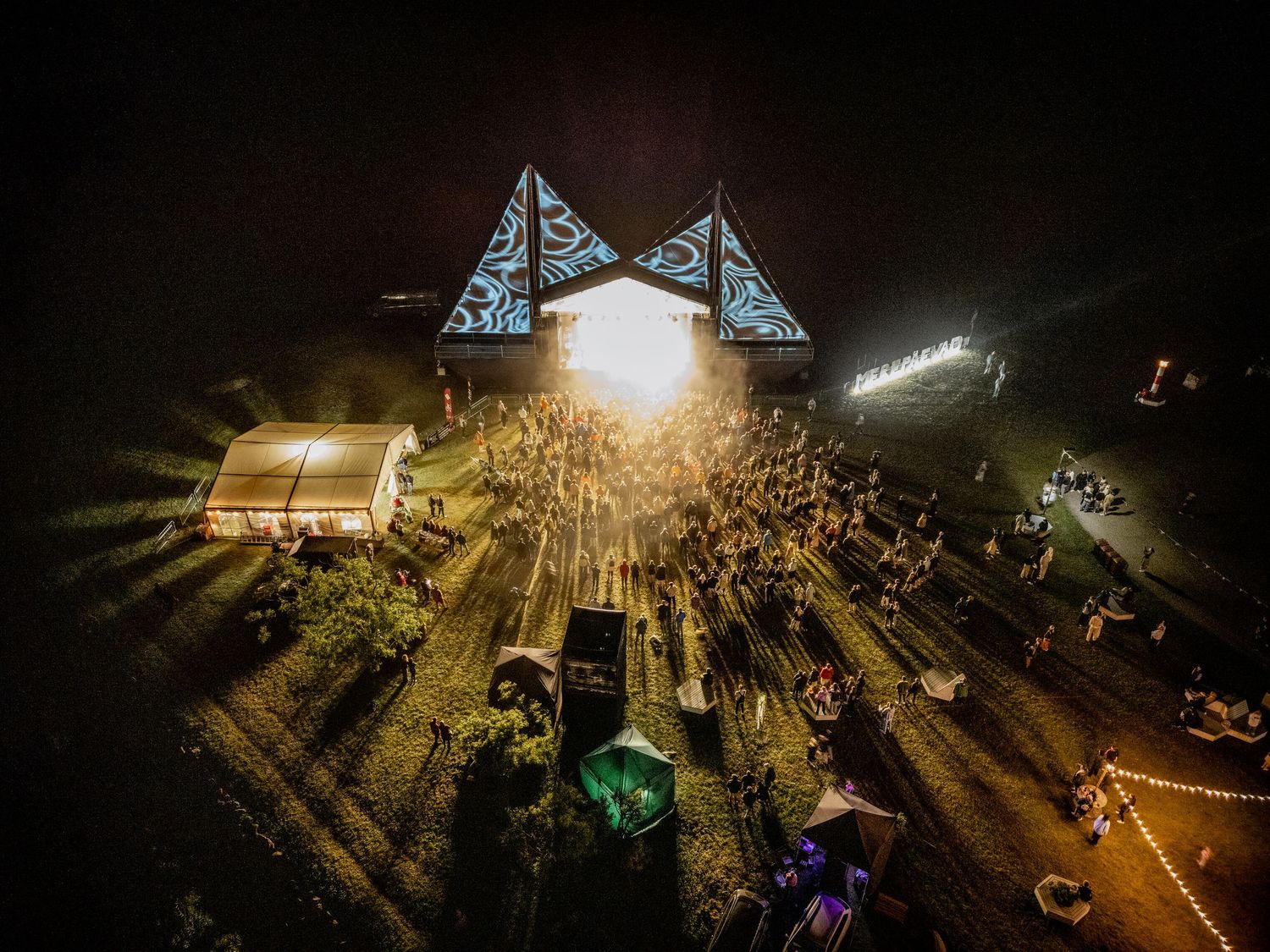 Aerial night view of a vibrant outdoor music festival with glowing stage triangles and crowds of people.