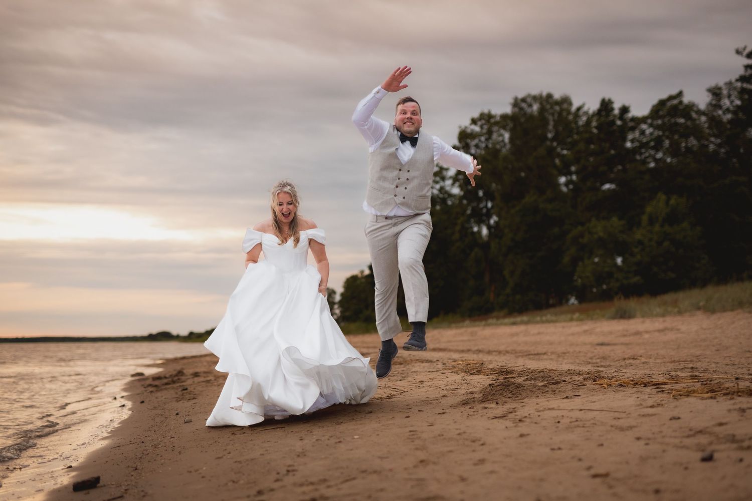Joyful newlyweds celebrate on a sandy beach, bride in white gown and groom jumping with excitement at sunset.