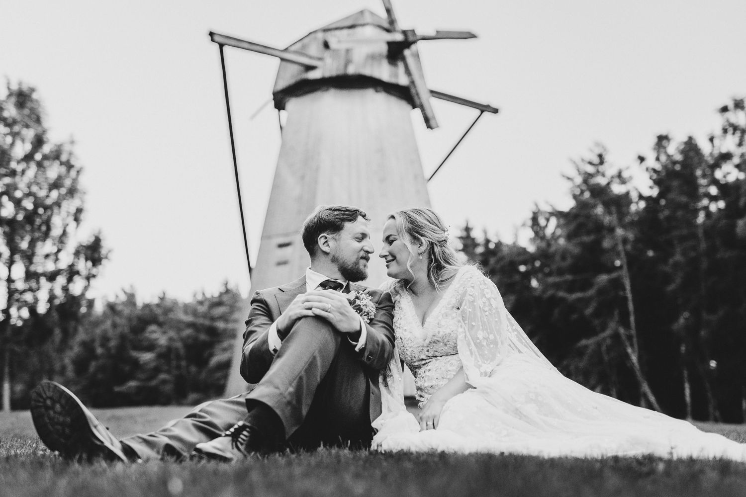Black and white photo of a bride and groom sitting on grass, gazing at each other with a windmill in the background.