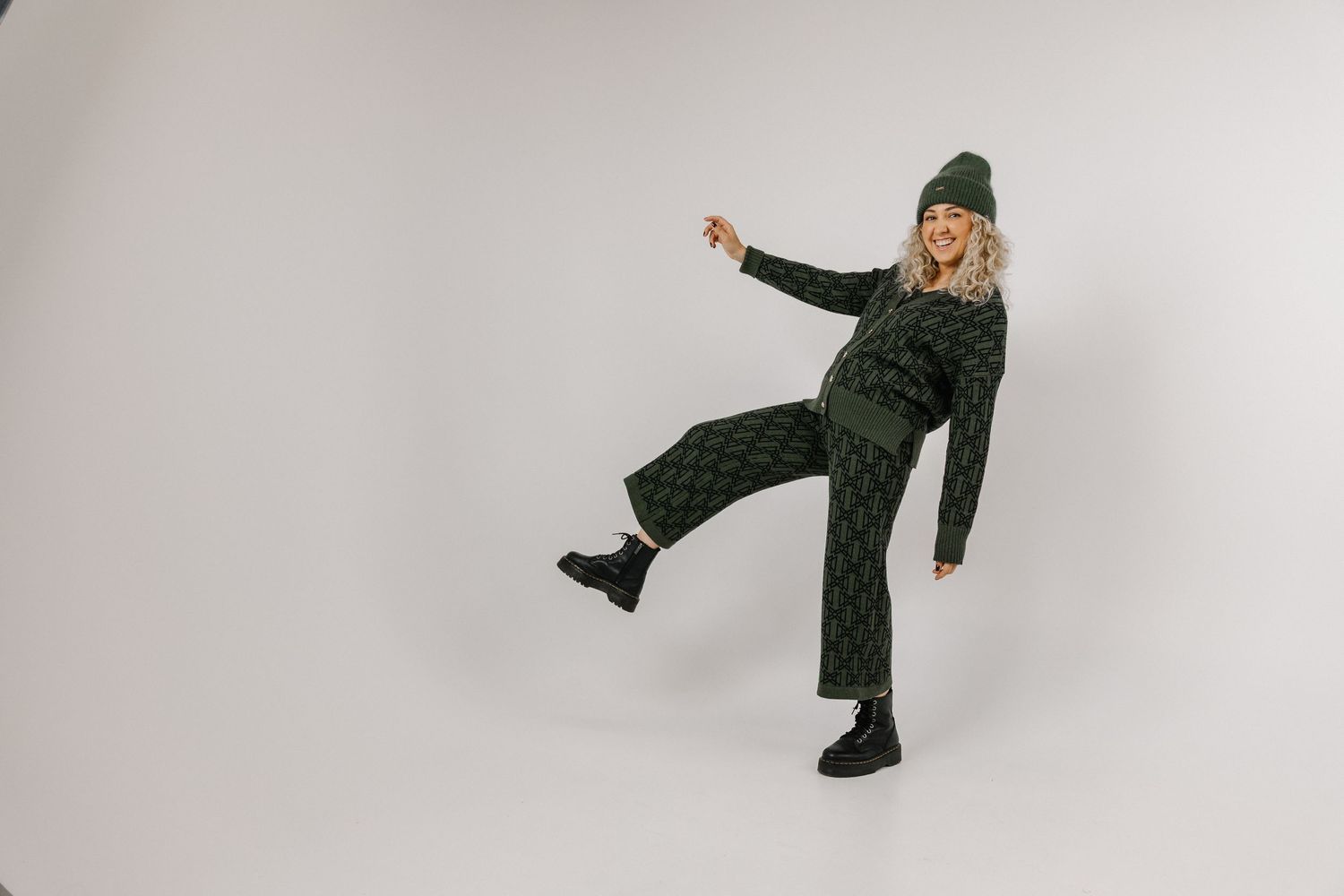 Woman in dark green knit matching set and beanie posing playfully with one leg raised on white background.