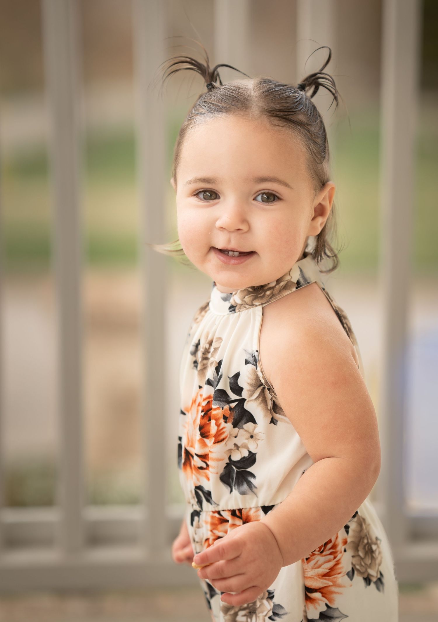 Adorable toddler girl in floral romper with tiny pigtails smiling outdoors.