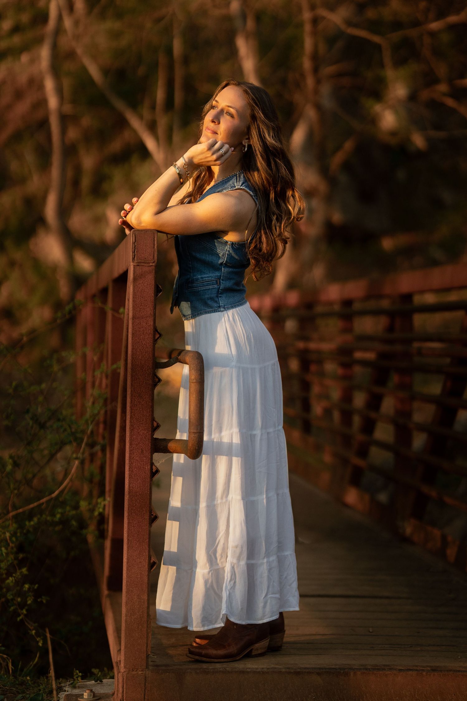 Young woman in denim vest and white maxi skirt posing on wooden bridge at sunset.