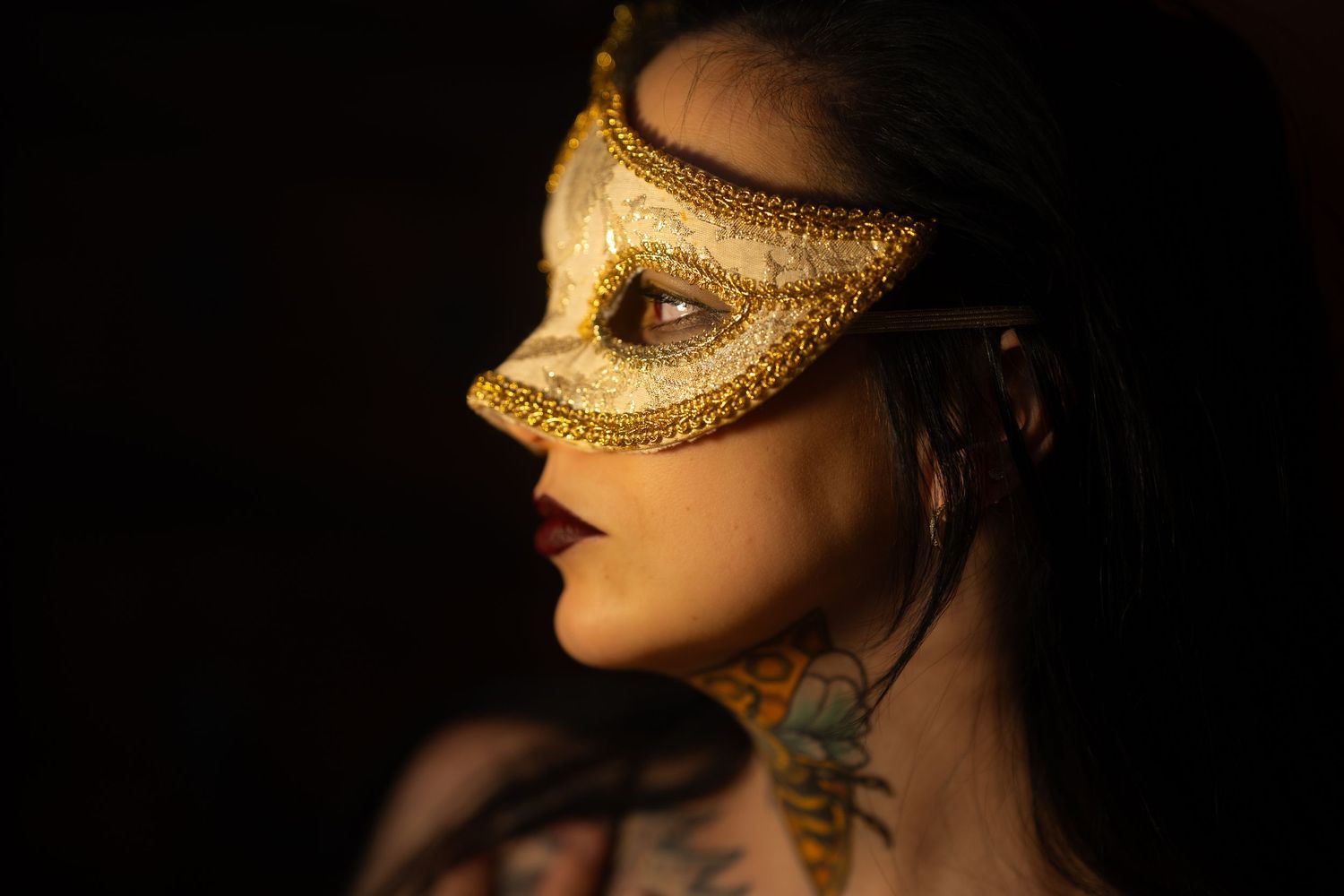 Woman wearing an ornate gold masquerade mask with tattoos on her neck, dark moody portrait.