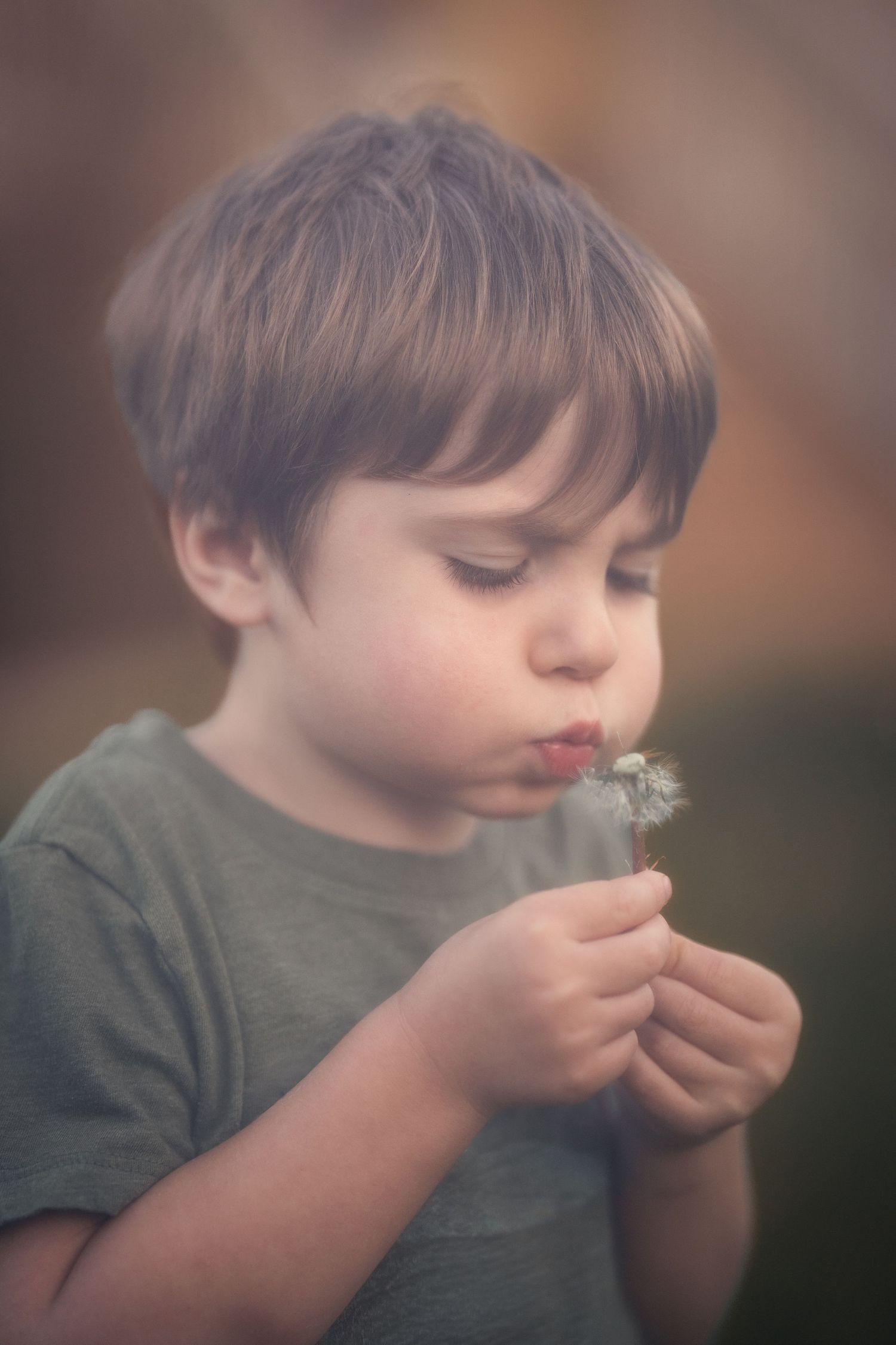 Young boy blowing dandelion seeds outdoors, wearing olive green shirt, close-up portrait with soft bokeh background.