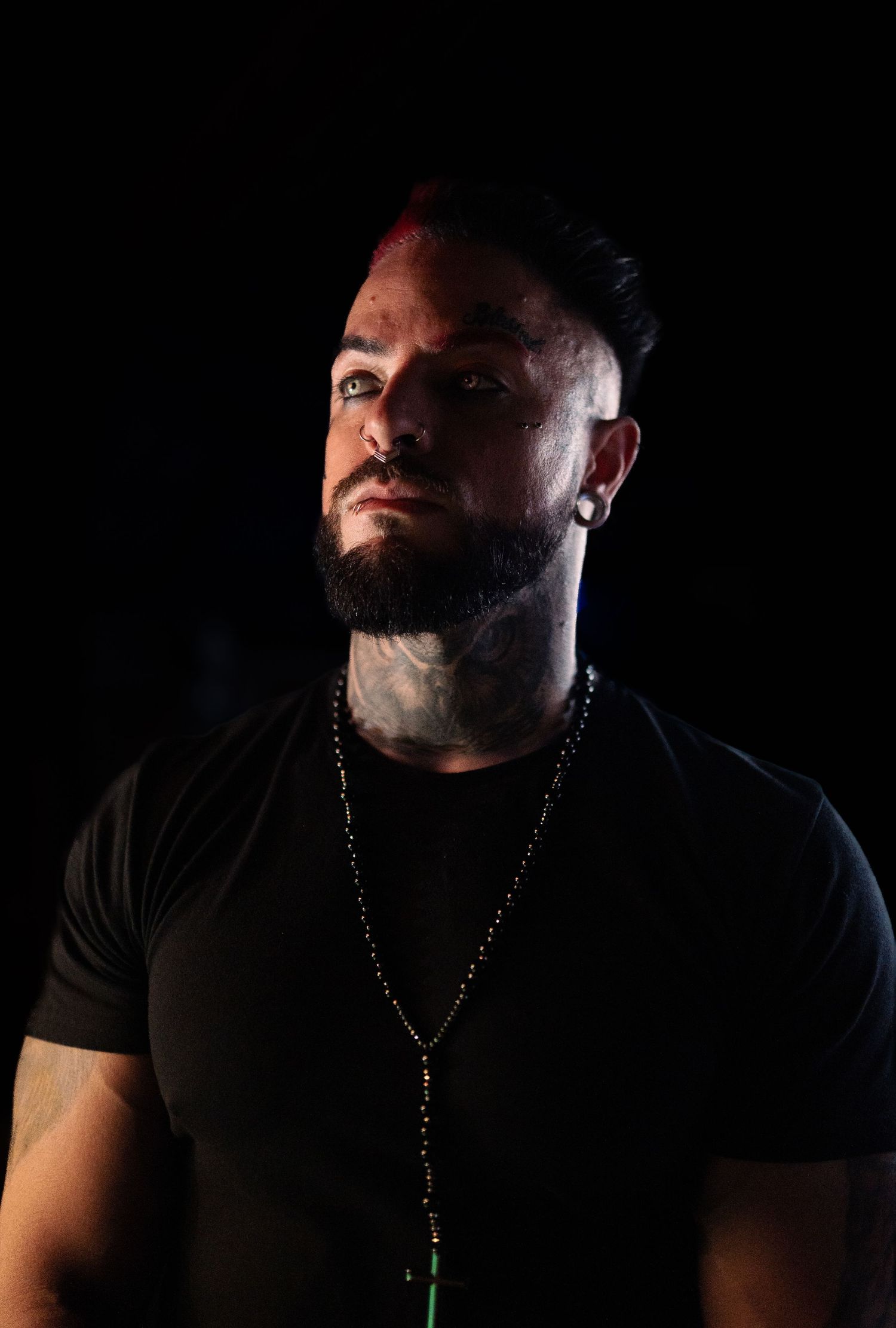 Tattooed man with beard wearing black t-shirt and beaded necklace posed against dark background.