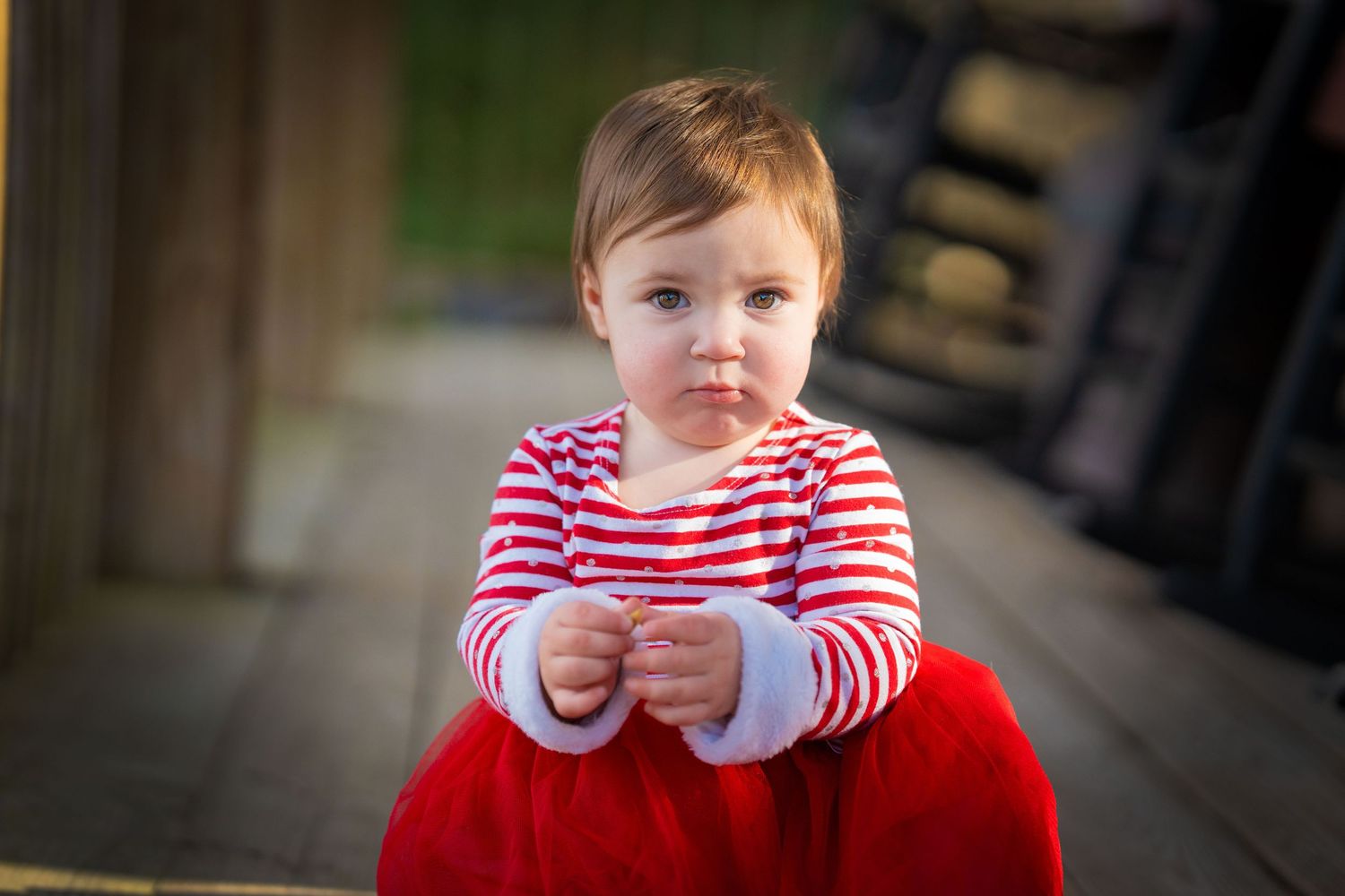 Adorable toddler girl in red skirt and striped shirt sitting on wooden deck.