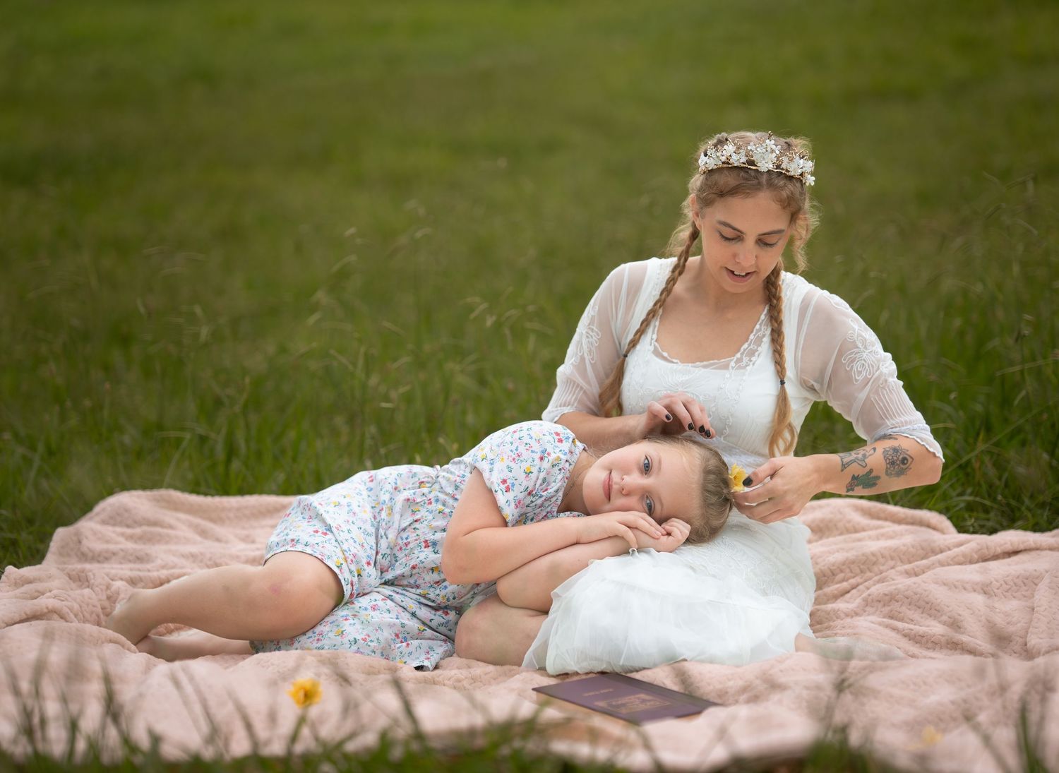 A mother braids her young daughter's hair on a pink blanket in a lush green meadow during a serene outdoor picnic.