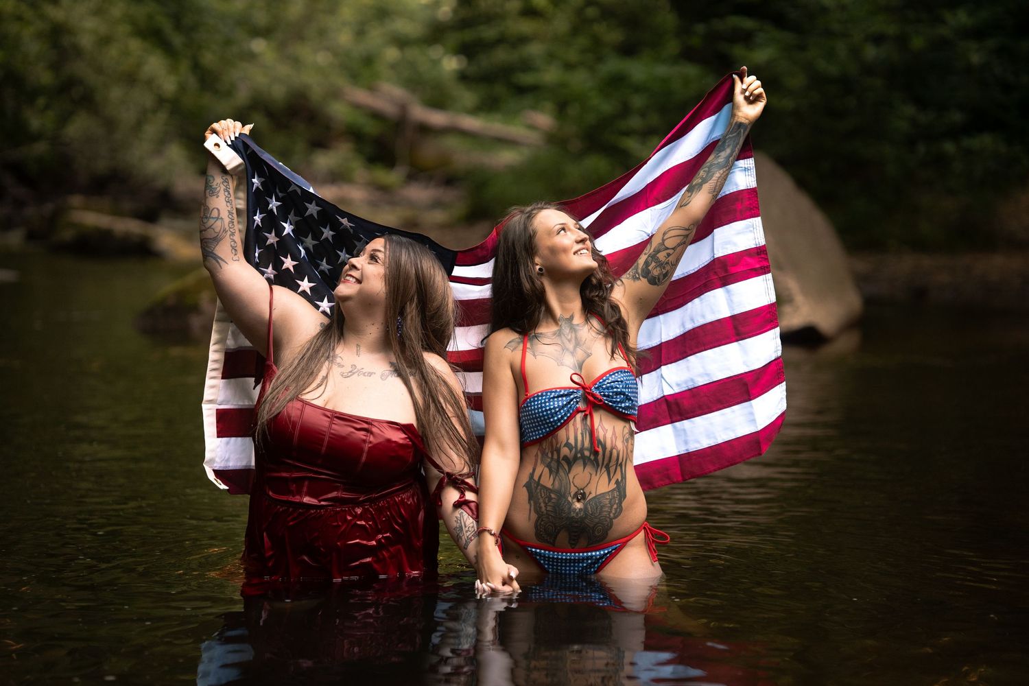 Two tattooed women holding an American flag while standing in a river, wearing patriotic swimwear.
