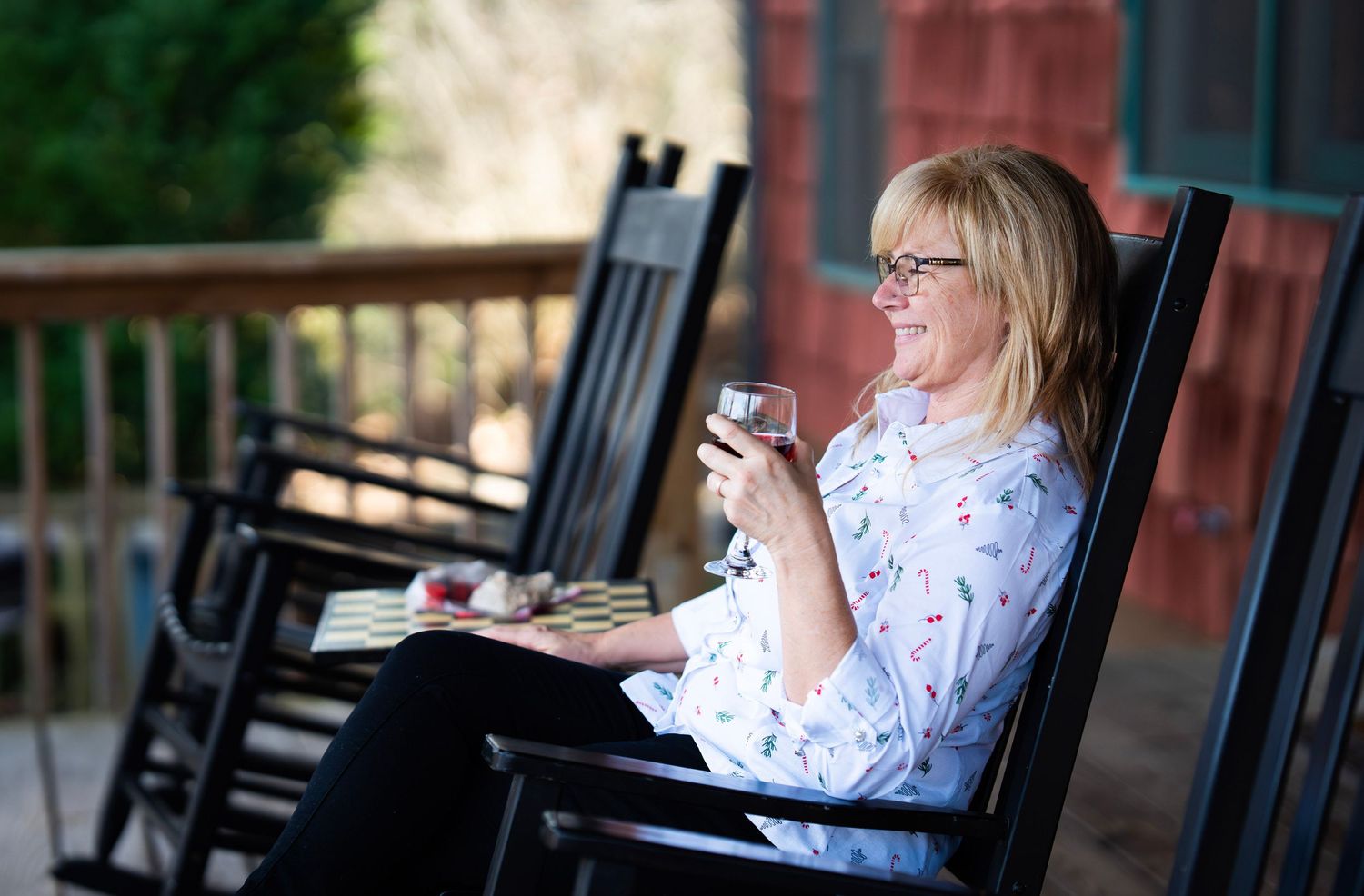 Smiling blonde woman enjoying wine in rocking chair on outdoor porch.