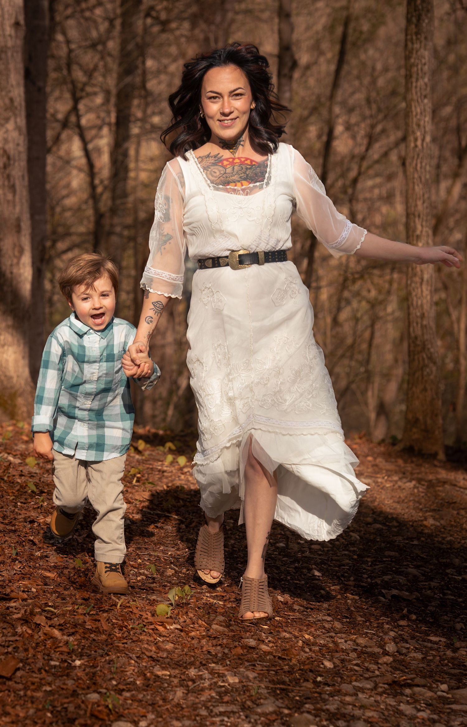 Woman in white dress and young boy holding hands running through autumn forest, smiling joyfully.