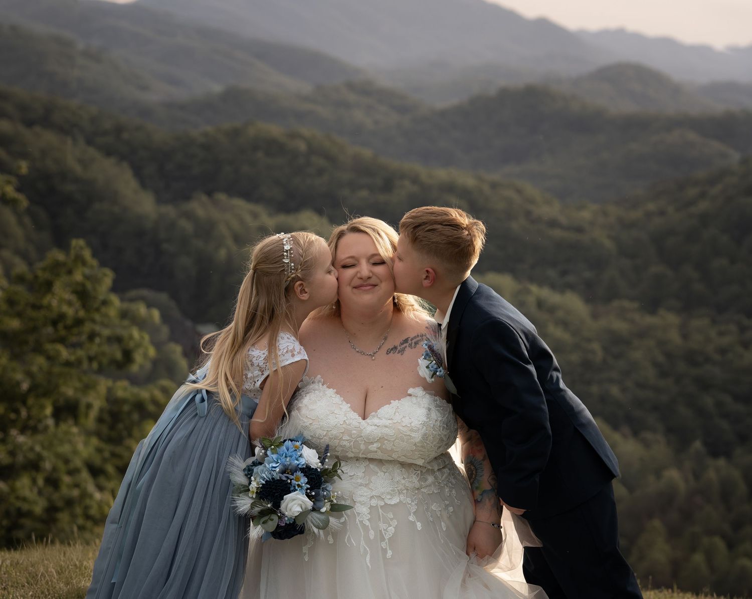 Bride in white lace gown holding blue bouquet receives kisses from children with mountain views behind.