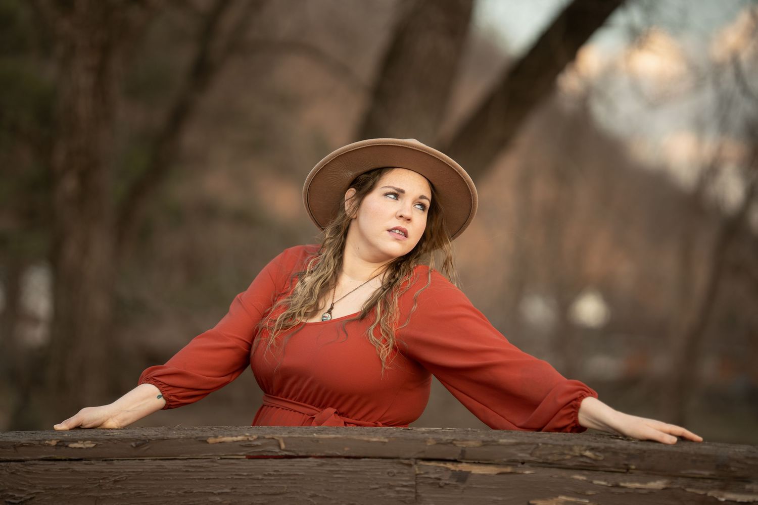 Woman in red dress and tan hat leans on wooden railing with arms spread wide.