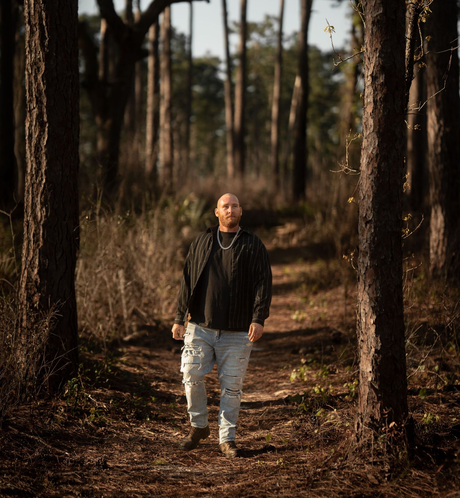 Bald man in black jacket and distressed jeans walking on forest trail between tall pine trees.
