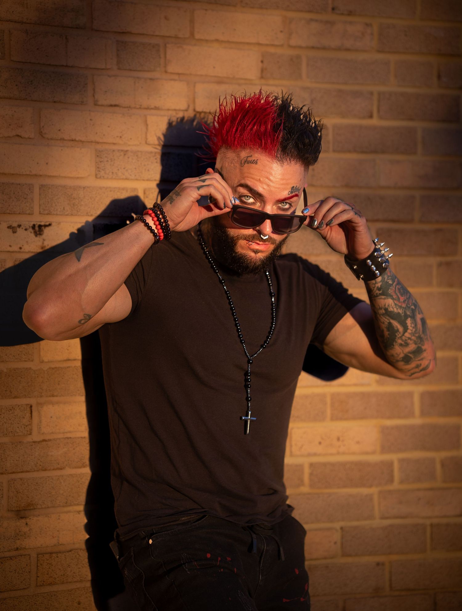 Tattooed man with colorful mohawk adjusting sunglasses wearing black t-shirt against brick wall.