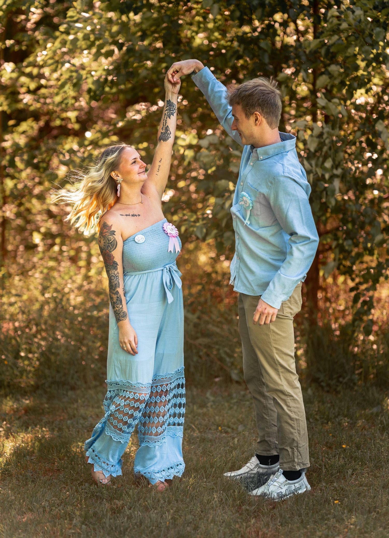 A couple dances outdoors in matching light blue outfits, surrounded by lush green trees in a sunlit garden setting.
