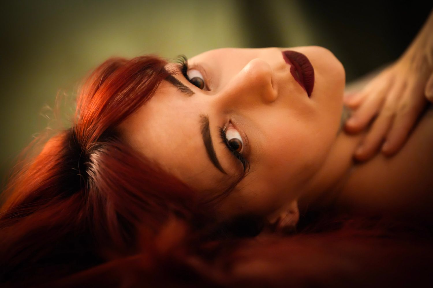 A woman with red hair and dark lipstick poses lying back with a serene expression in warm lighting.