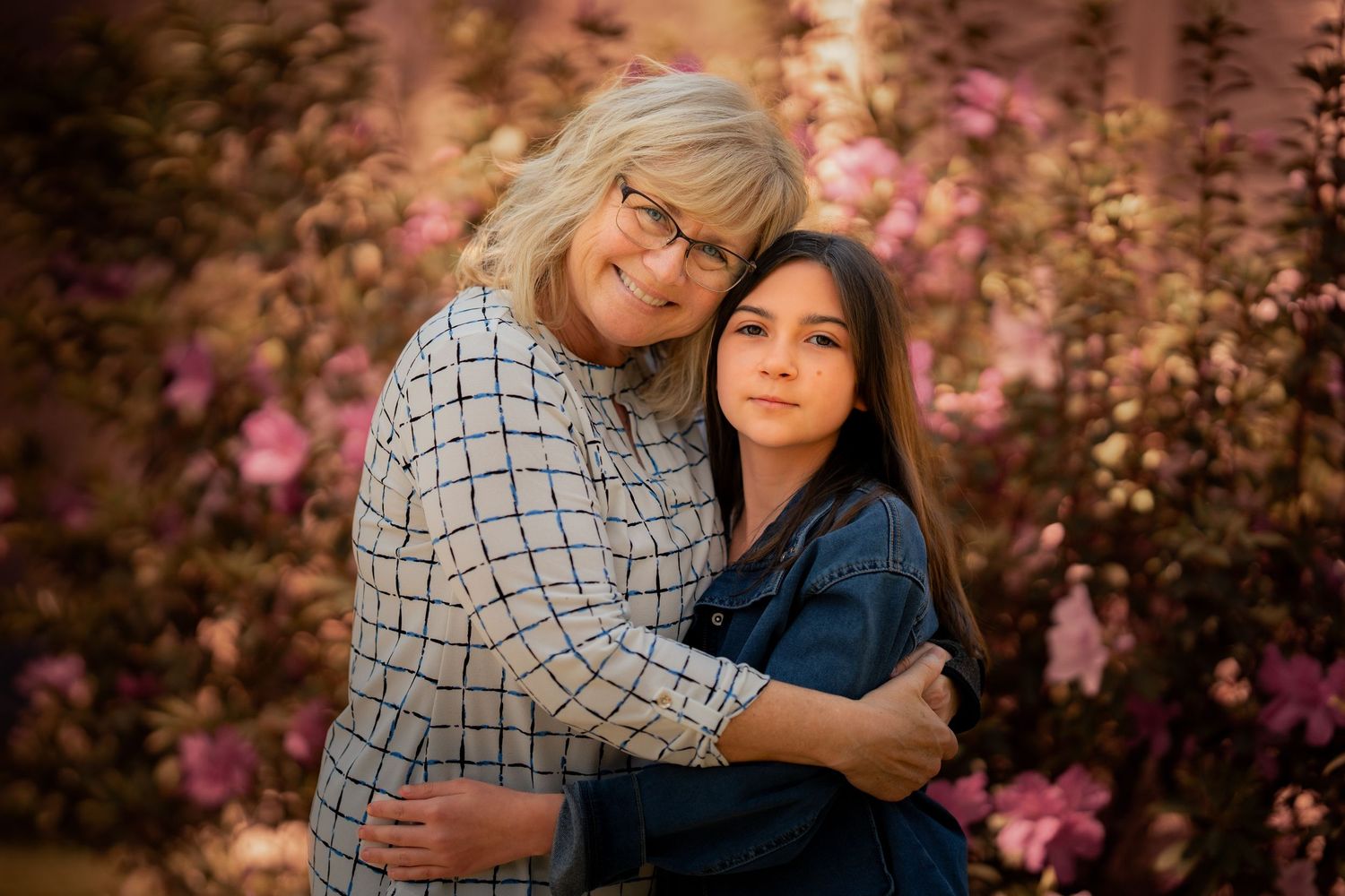 Grandmother hugging young dark-haired girl in front of pink flowering bushes.