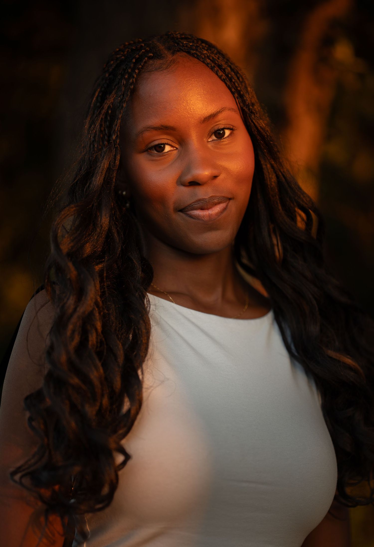 Woman with long braided hair wearing a white top posing in warm evening light.