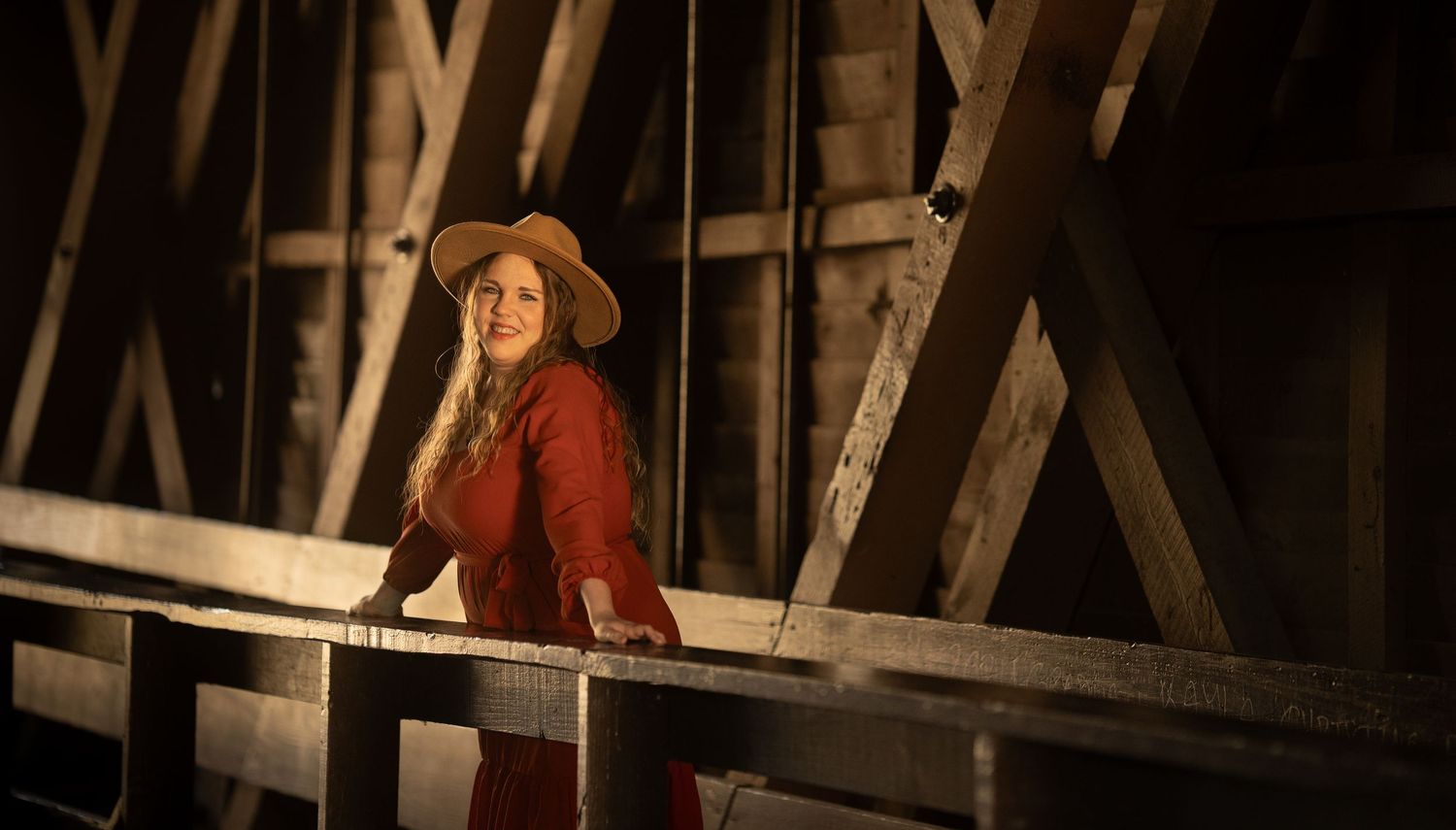 Woman in red dress and straw hat stands inside rustic wooden covered bridge.