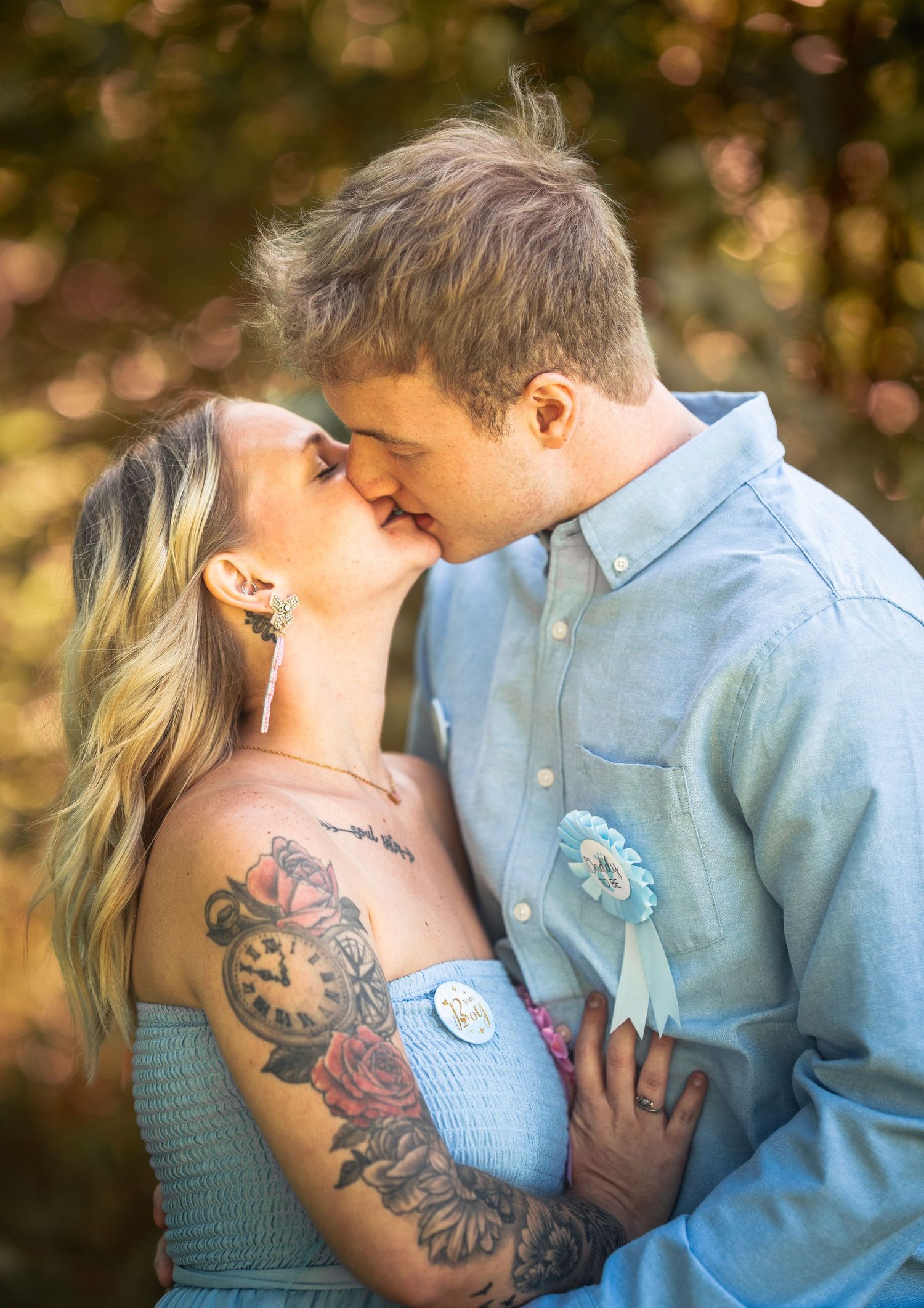A couple sharing a romantic kiss outdoors, woman with floral tattoos in blue dress, man in light blue button-up shirt.