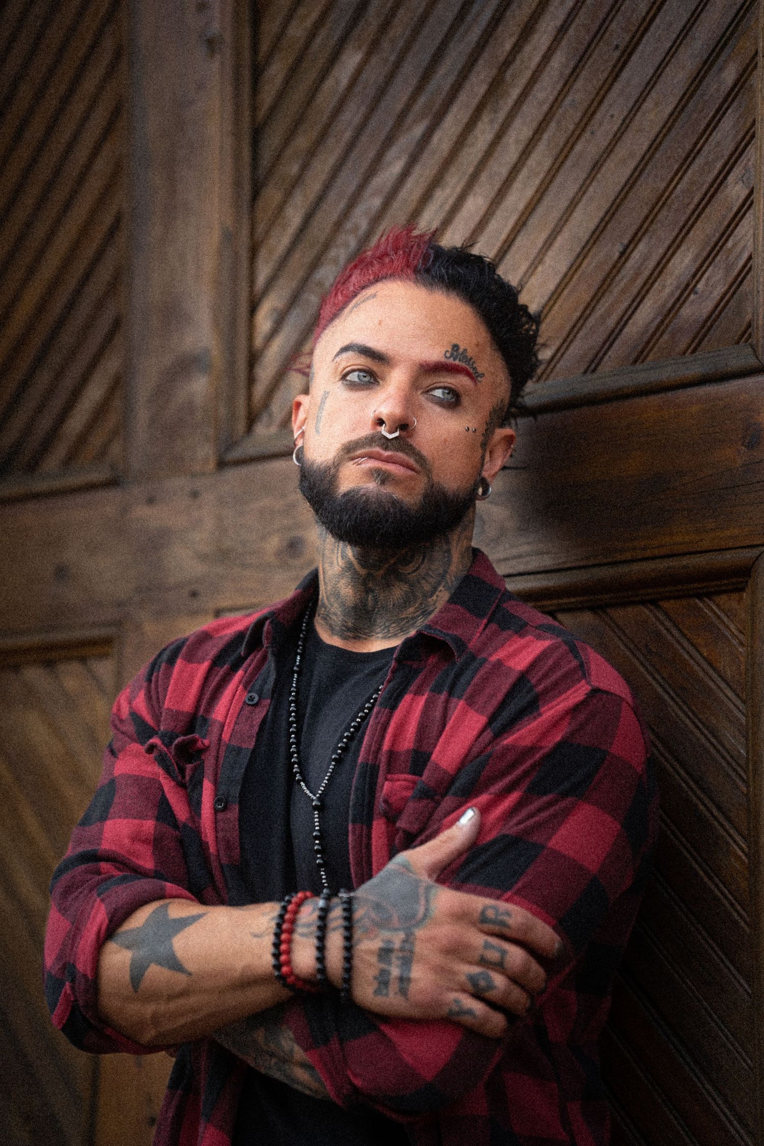 Tattooed man with red mohawk wearing plaid shirt standing arms crossed against wooden wall.