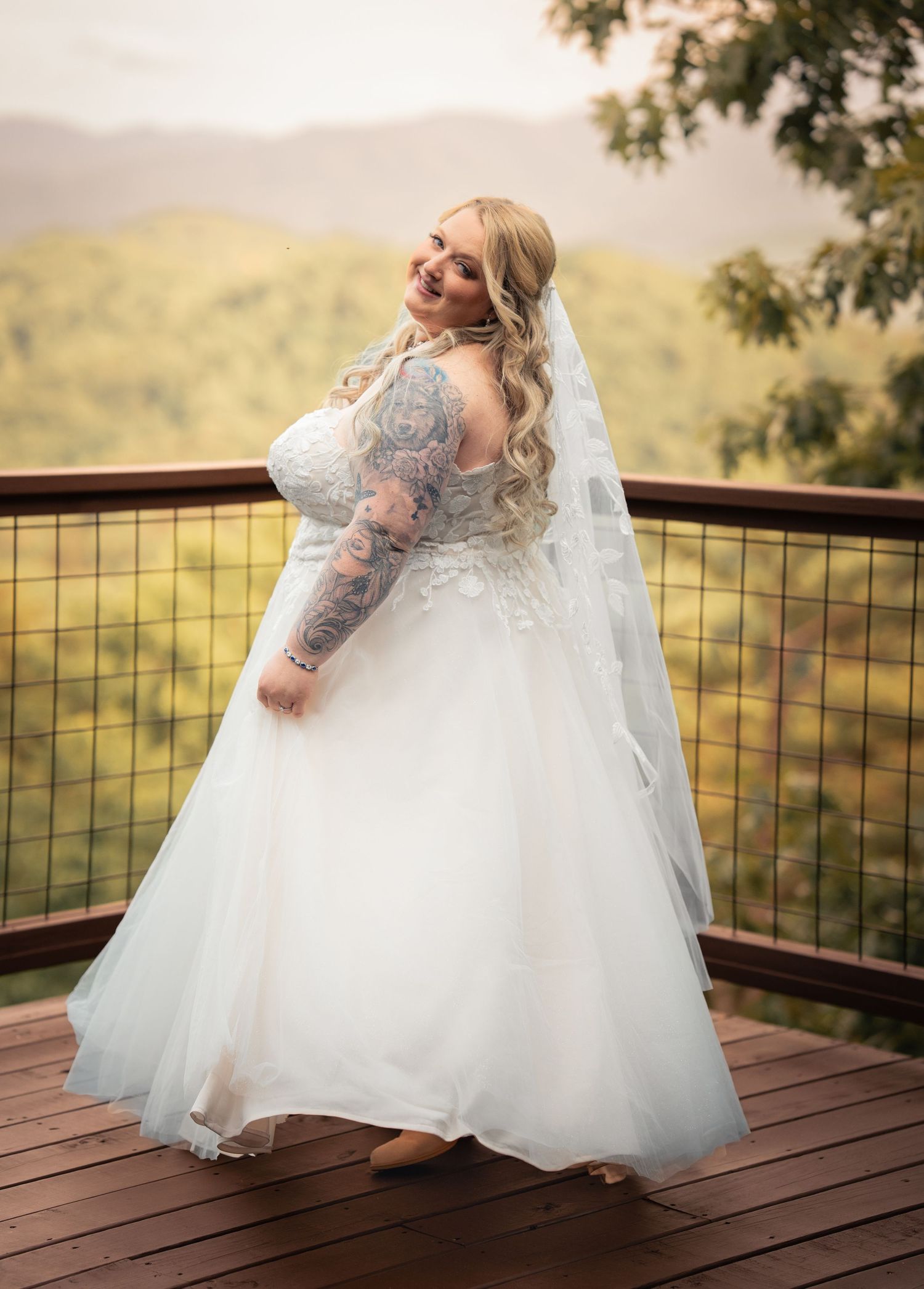 Bride in a white lace wedding gown with tattoo sleeves and veil poses on a deck with mountain views.