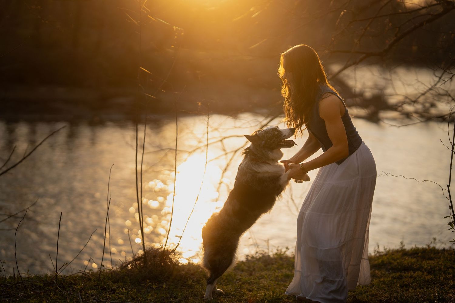 Woman in white dress playing with jumping dog near reflective lake at golden sunset.