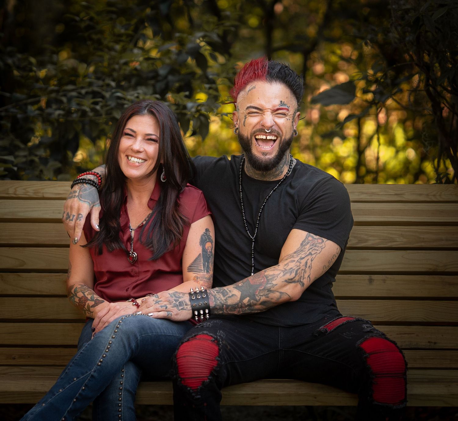 Smiling tattooed couple embracing on a park bench surrounded by autumn foliage.