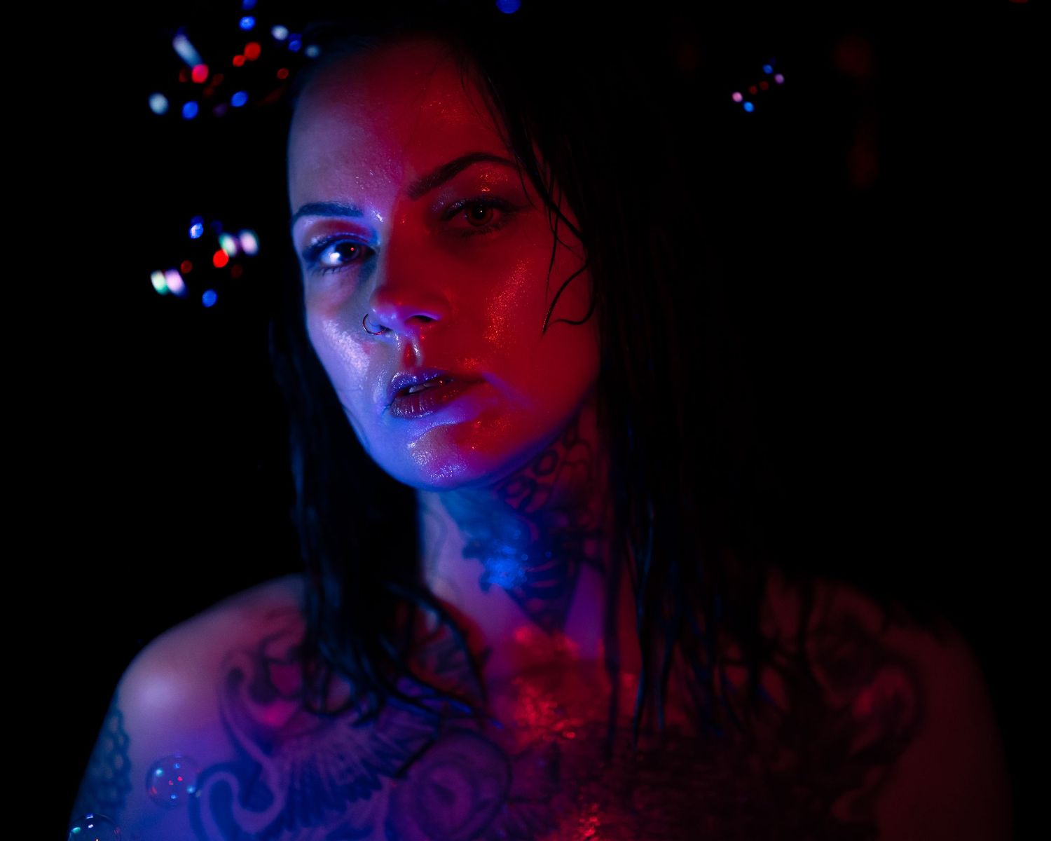Woman with neck tattoos and body art posed under dramatic red and blue neon lighting in dark setting.