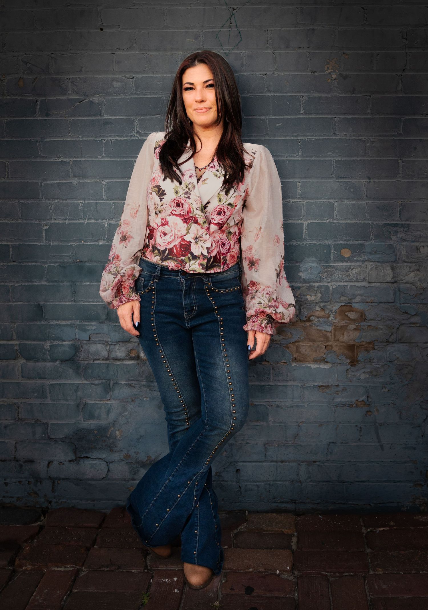 Woman in floral blouse and studded bell-bottom jeans poses against gray brick wall.