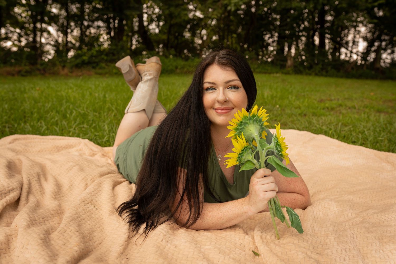 Young woman with dark hair lying on a blanket outdoors, holding sunflowers, smiling in a green field.