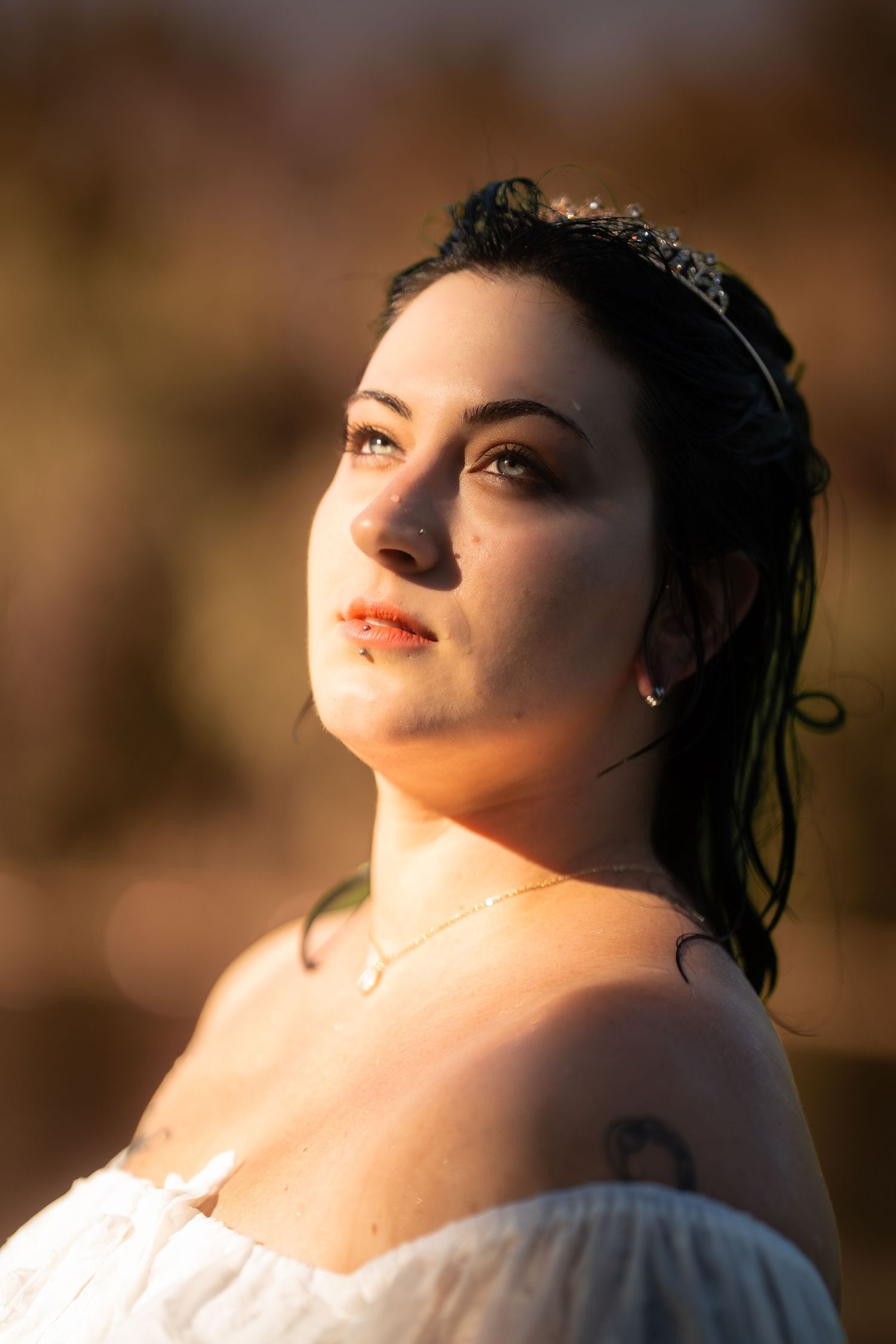 Woman wearing a tiara and white dress with a tattoo posing in warm golden sunlight.