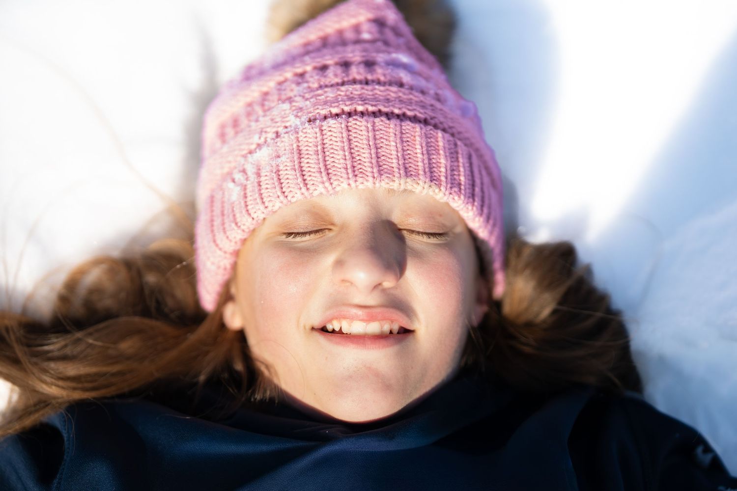 Young girl in pink knit hat lying in snow with eyes closed smiling peacefully.