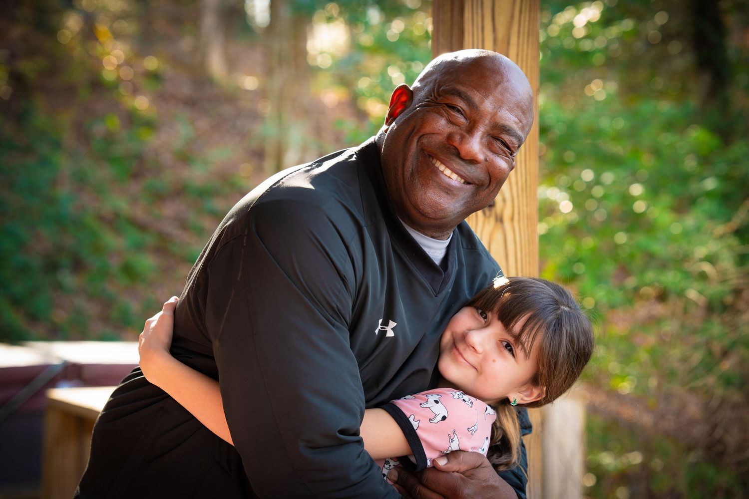 Smiling bald man hugging happy young girl outdoors near wooden post.